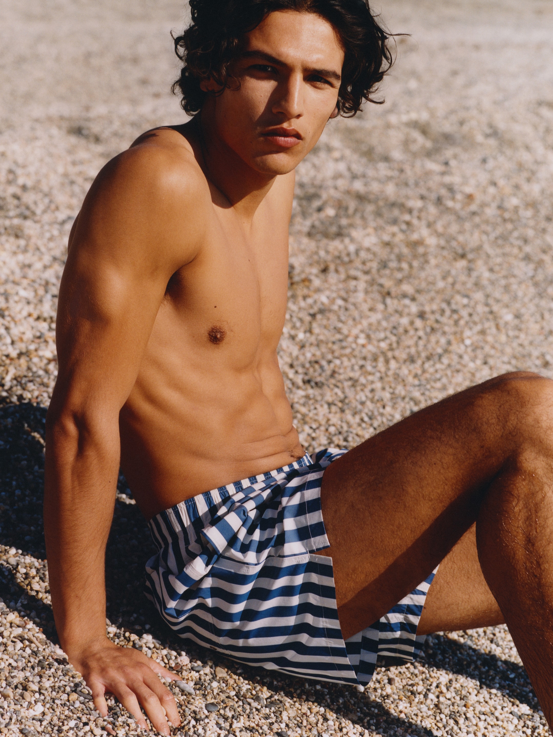 Shirtless person sitting on a pebbled beach, wearing striped shorts, with a neutral expression, and sunlight casting shadows on their skin.