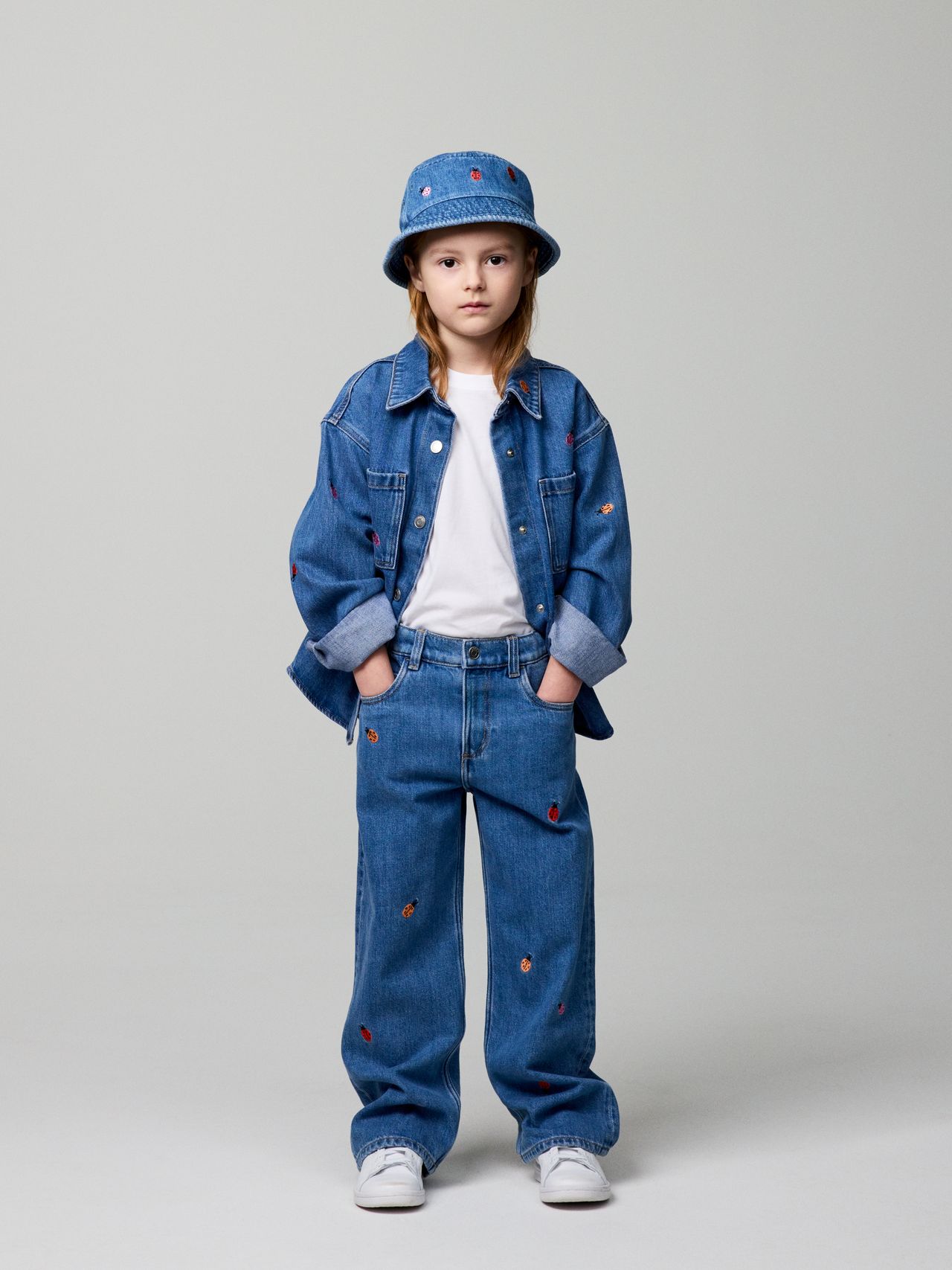 Child wearing a blue denim outfit with red accents, including a jacket, pants, and matching hat, standing against a plain background.