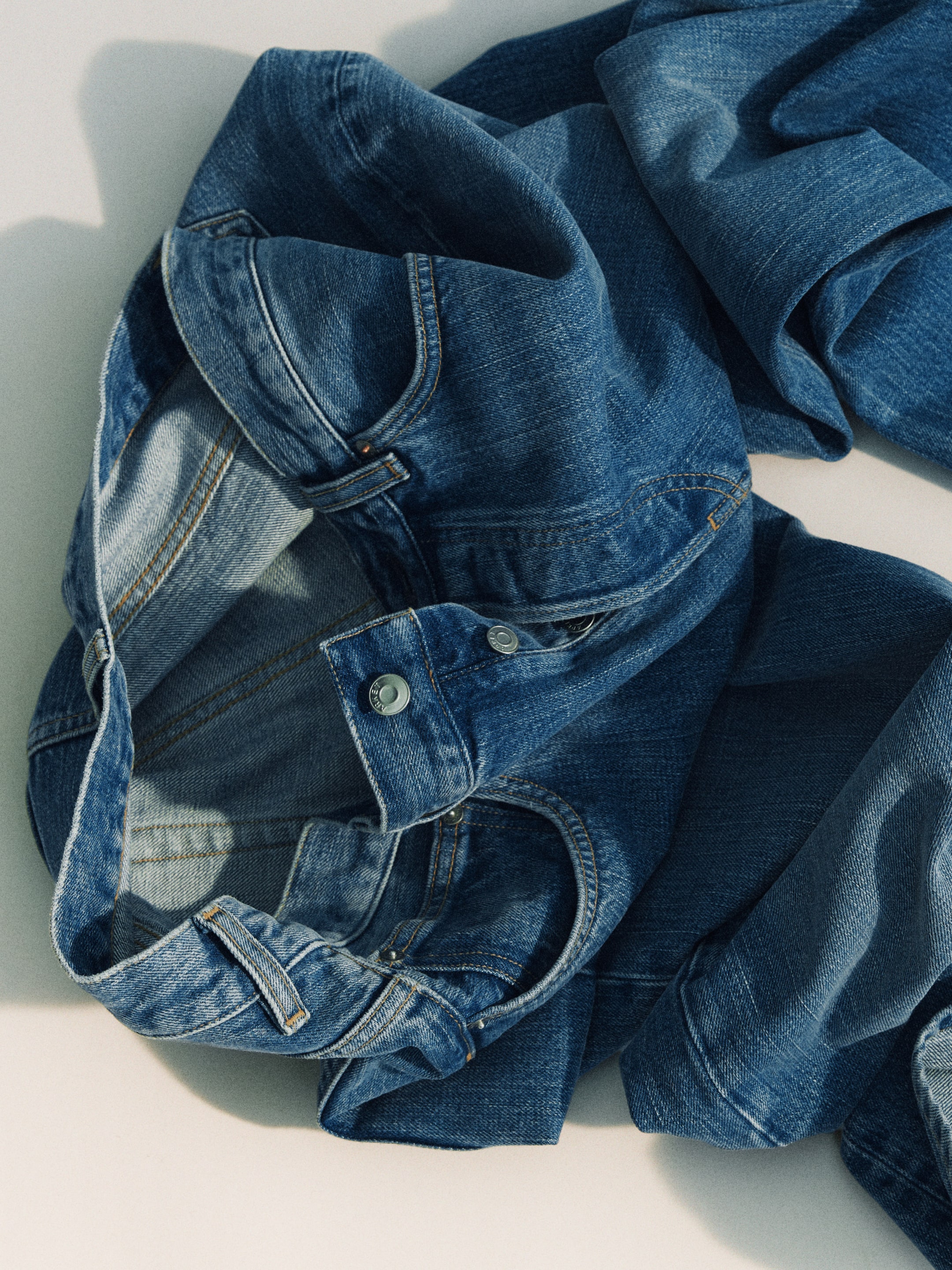 A crumpled pair of blue denim jeans lies on a light surface, showcasing detailed stitching and a metal button.