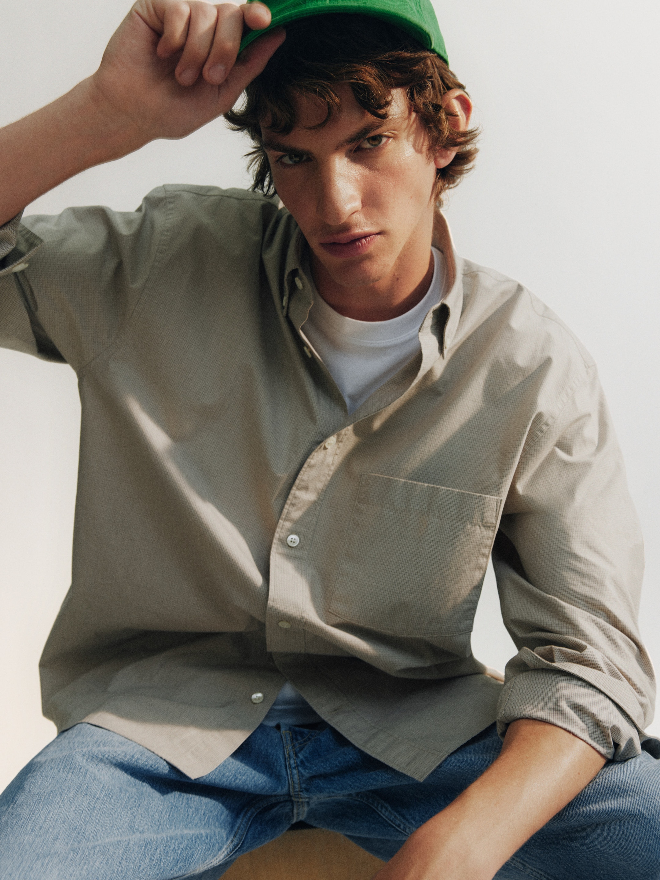 Young man in a beige shirt and jeans, tipping a green hat with one hand, looking into the camera against a neutral background.