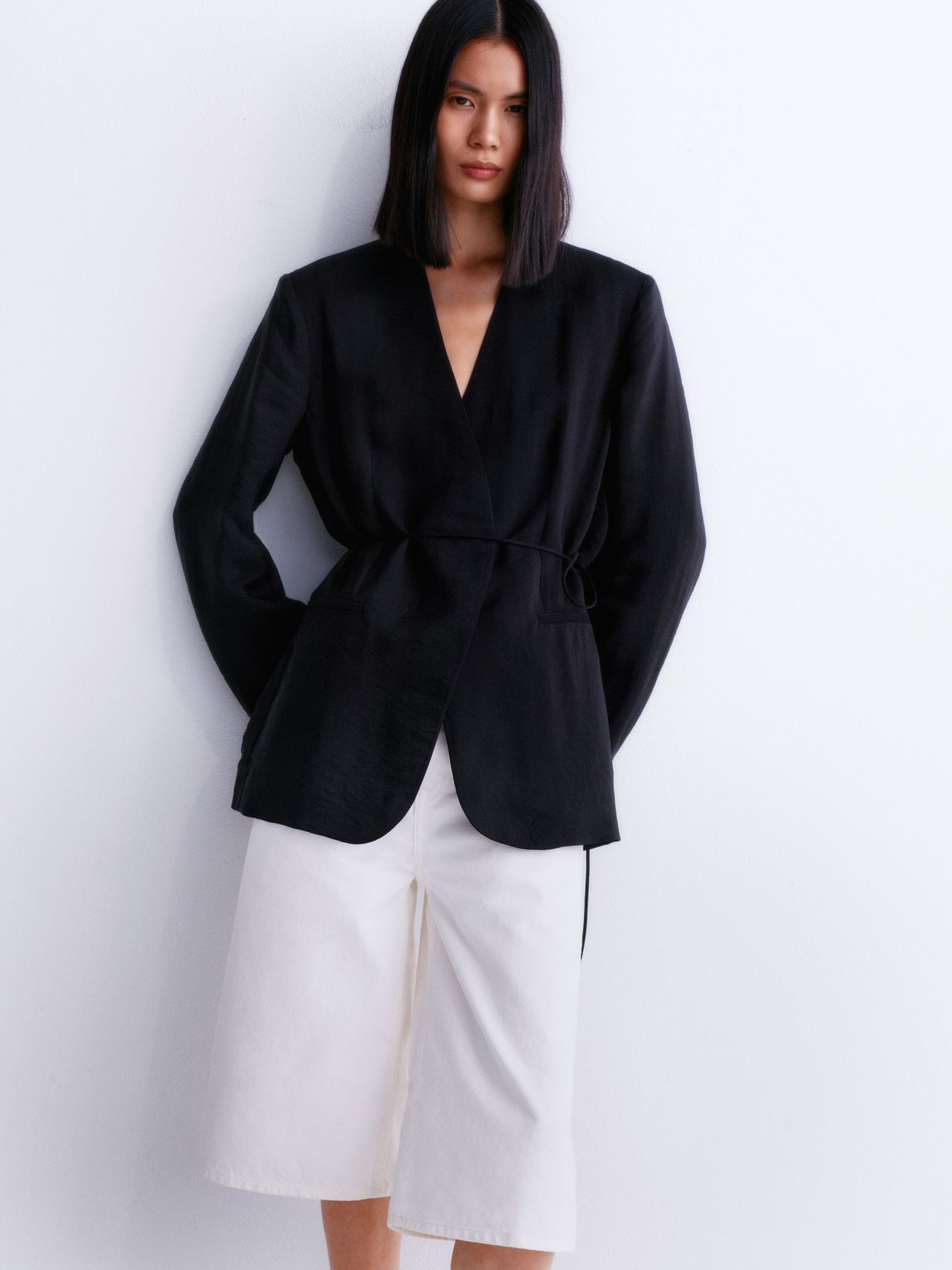 Person in a black V-neck blazer and white knee-length shorts against a plain white background, looking at the camera.