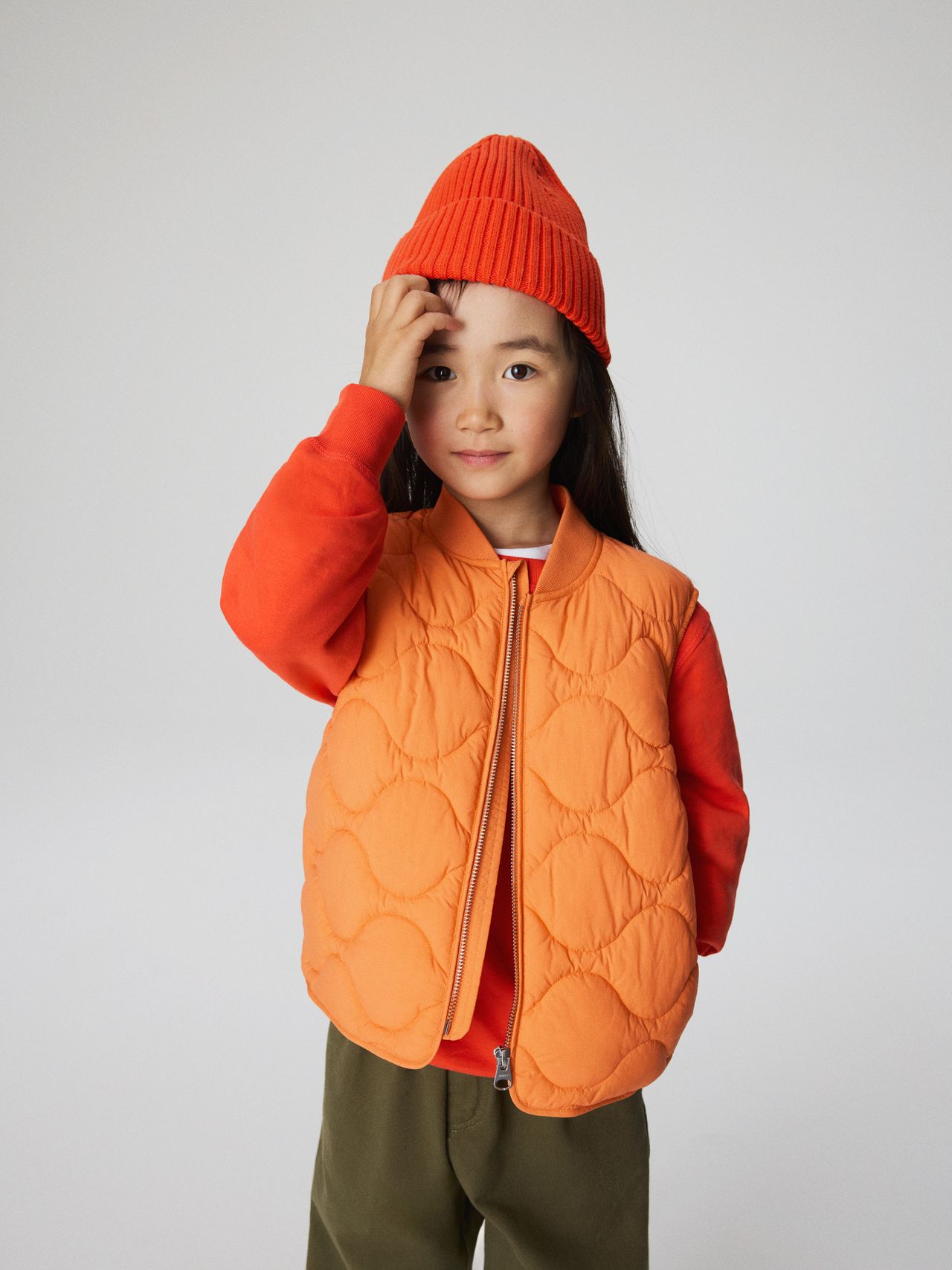 A child wearing an orange beanie and quilted vest over a red sweater, with olive green pants, stands against a plain white background.