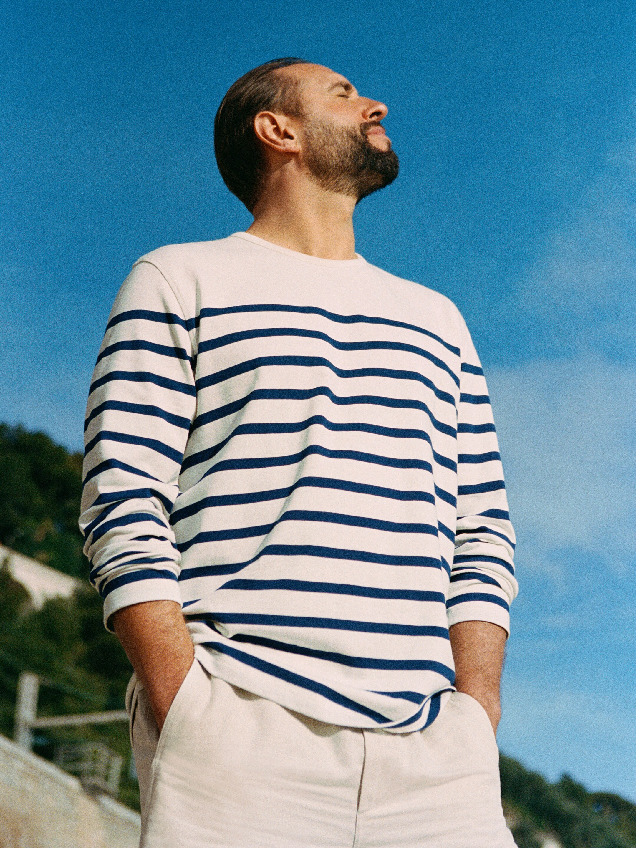 A man in a striped shirt and light pants stands outdoors, looking up at the sky with hands in pockets, against a backdrop of trees and blue sky.