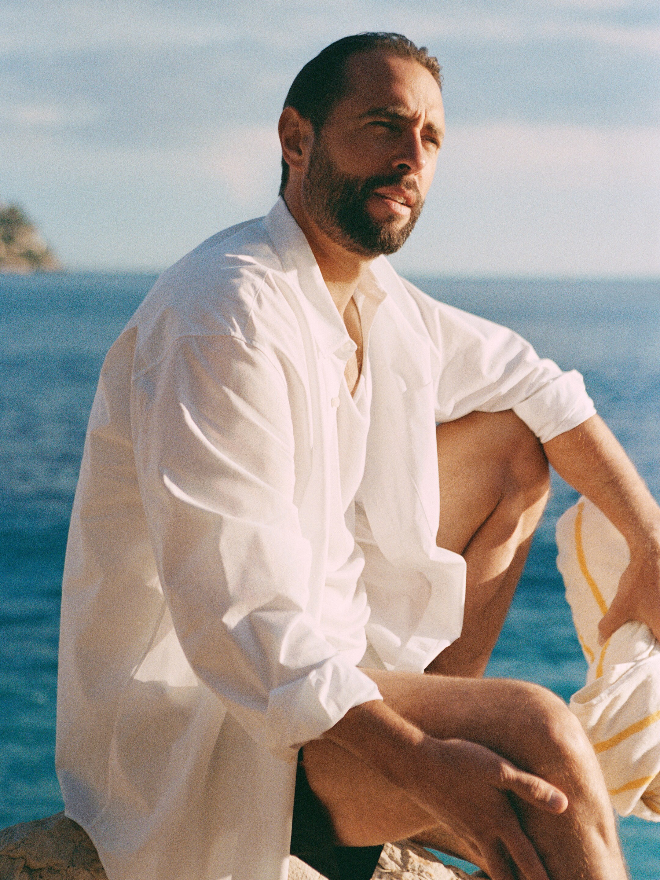 A man in a white shirt sits on a rock by the ocean, gazing into the distance, with a towel draped over his knee.