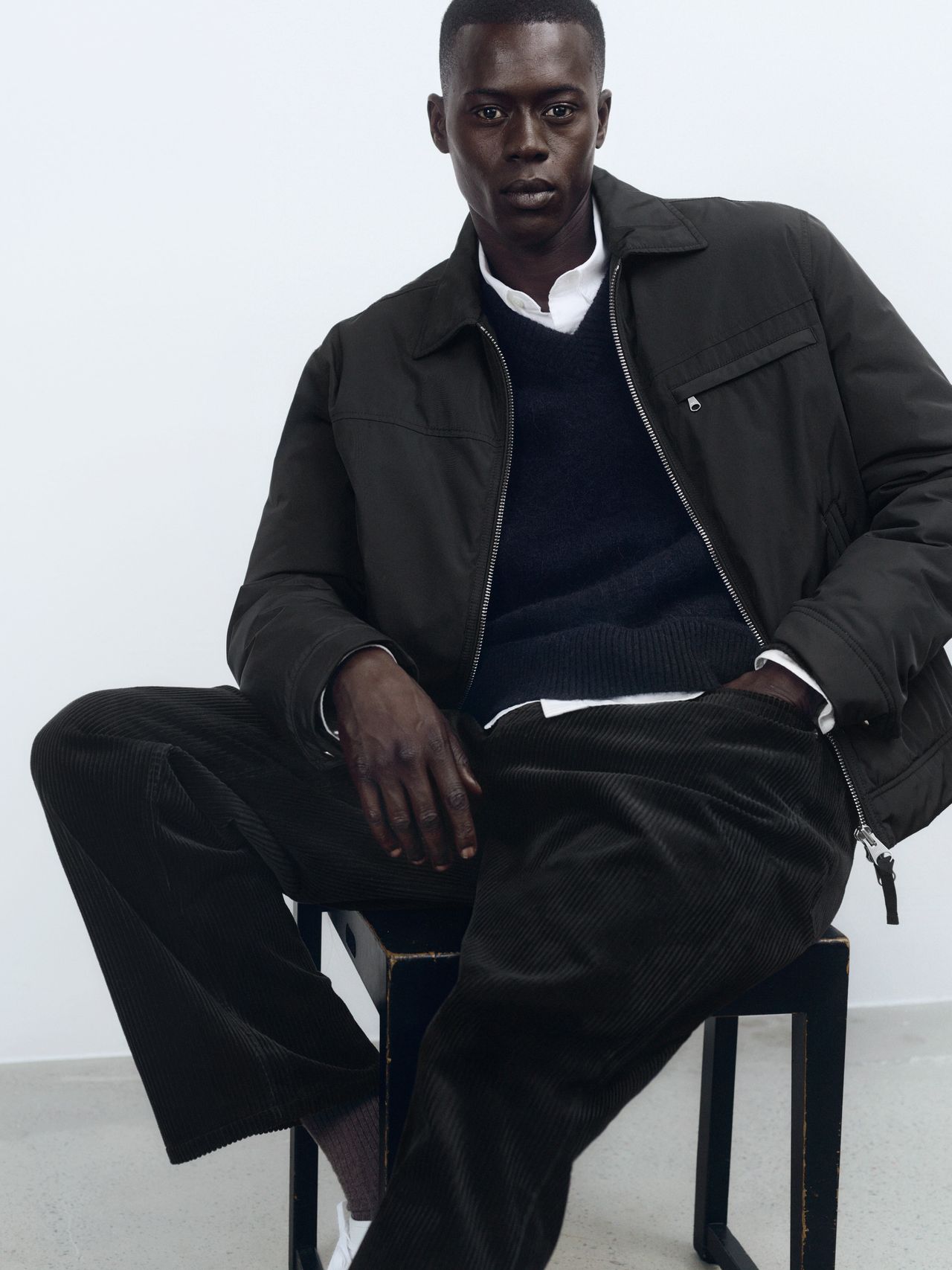 Man seated, wearing a dark jacket, sweater, and pants, with a white shirt. He is posing against a plain white background.
