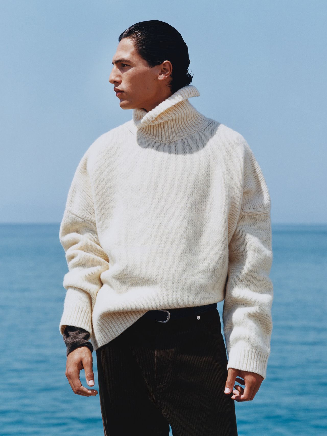 Person in a cream turtleneck sweater stands by the sea, looking to the side. Clear blue sky and ocean in the background.