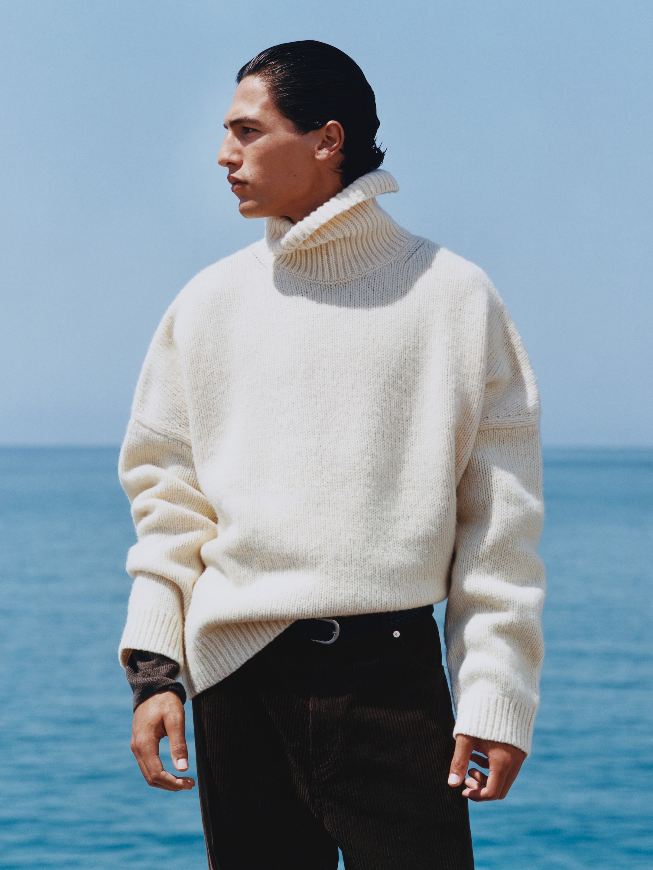 Person in a cream turtleneck sweater stands by the sea, looking to the side. Clear blue sky and ocean in the background.