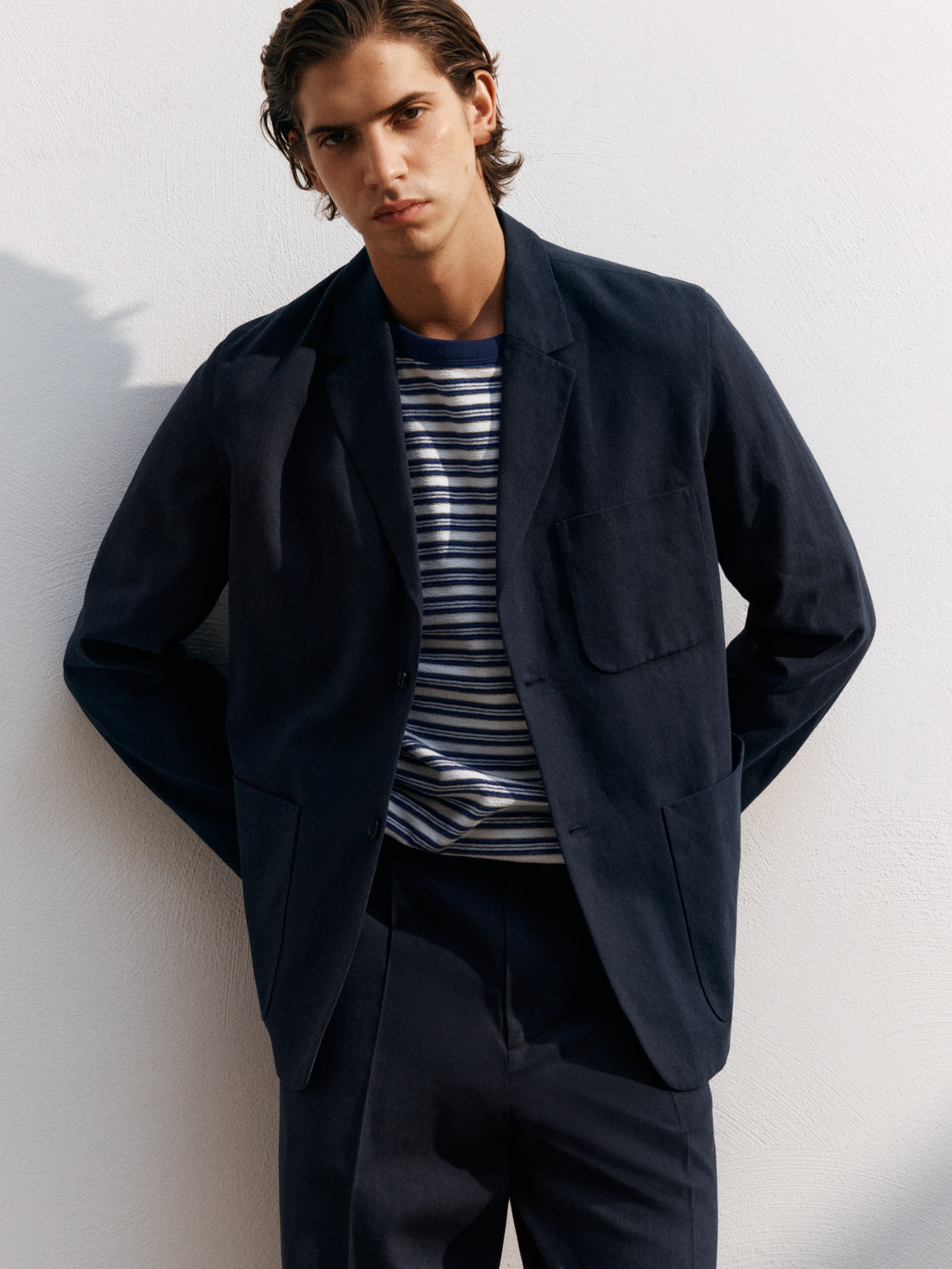 Person in a navy suit and striped shirt poses against a white wall, hands in pockets, casting a shadow.