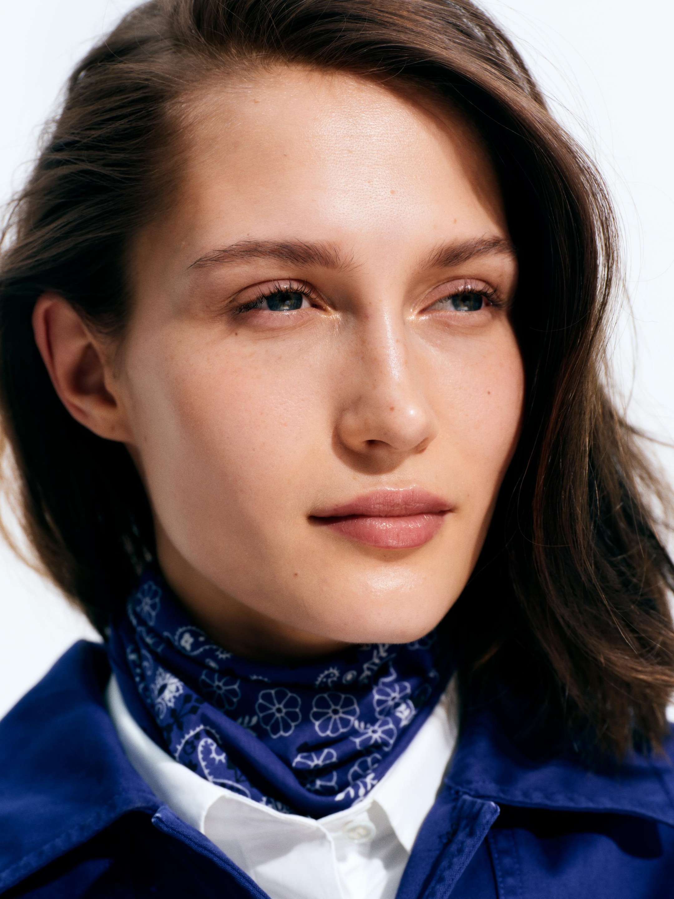A person with long brown hair wearing a blue scarf with floral patterns and a blue jacket, gazing thoughtfully to the side.