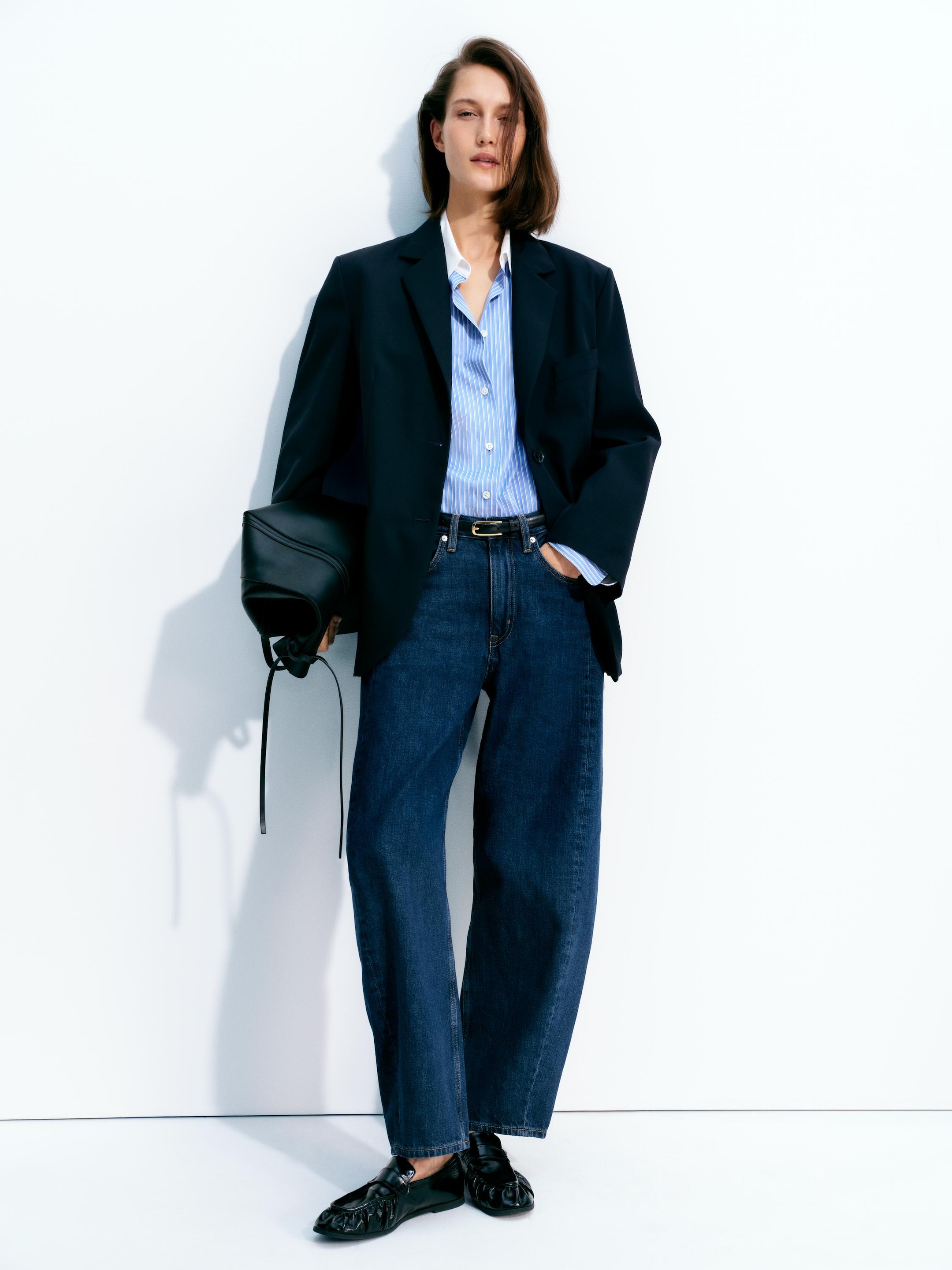 Person wearing a black blazer, blue striped shirt, and jeans, holding a black bag against a white background.