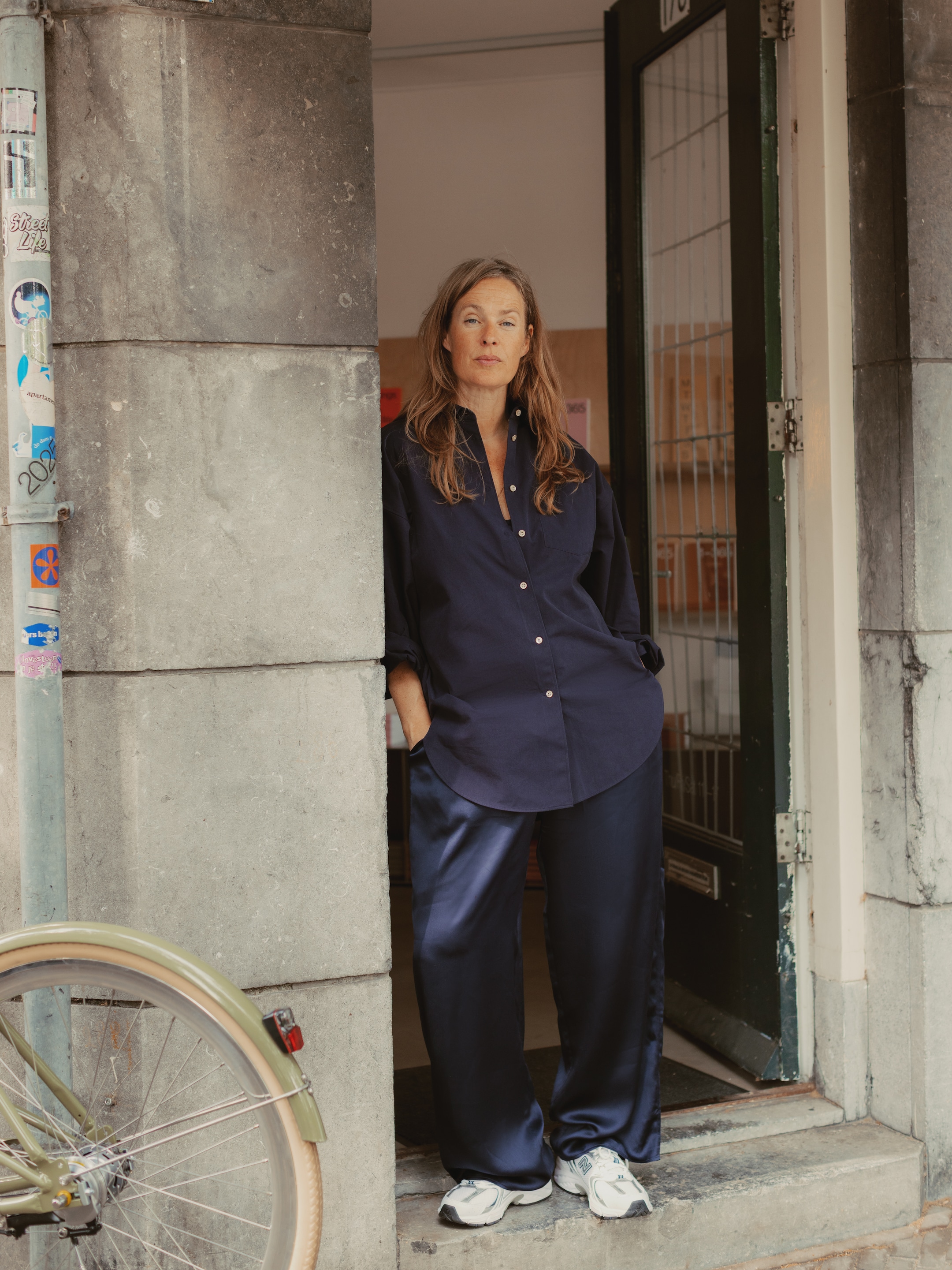 A person in a navy outfit stands in a doorway, leaning against a stone wall. A bicycle wheel is partially visible in the foreground.
