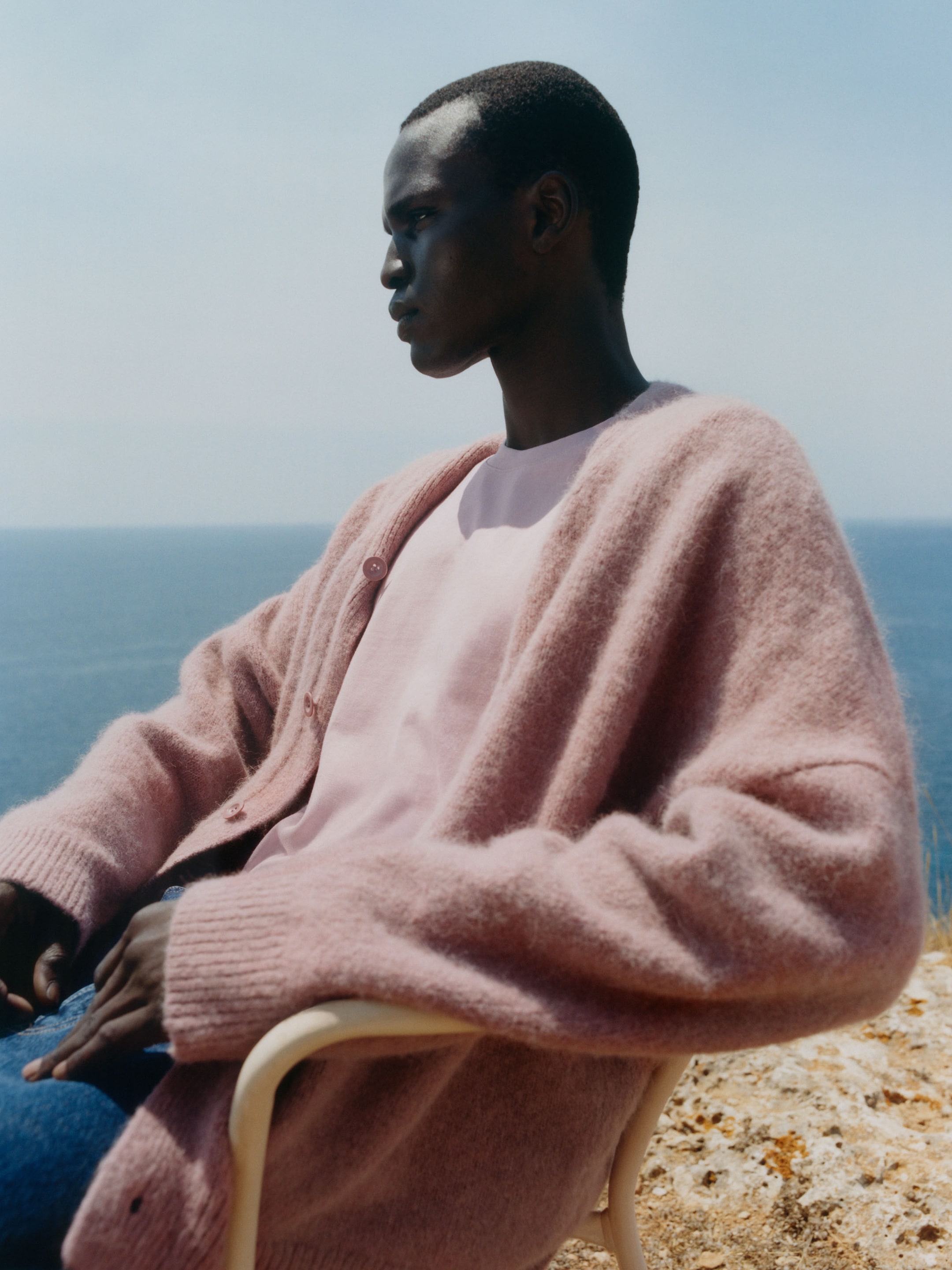A person in a pink cardigan sits on a chair, gazing thoughtfully out to sea against a clear blue sky.