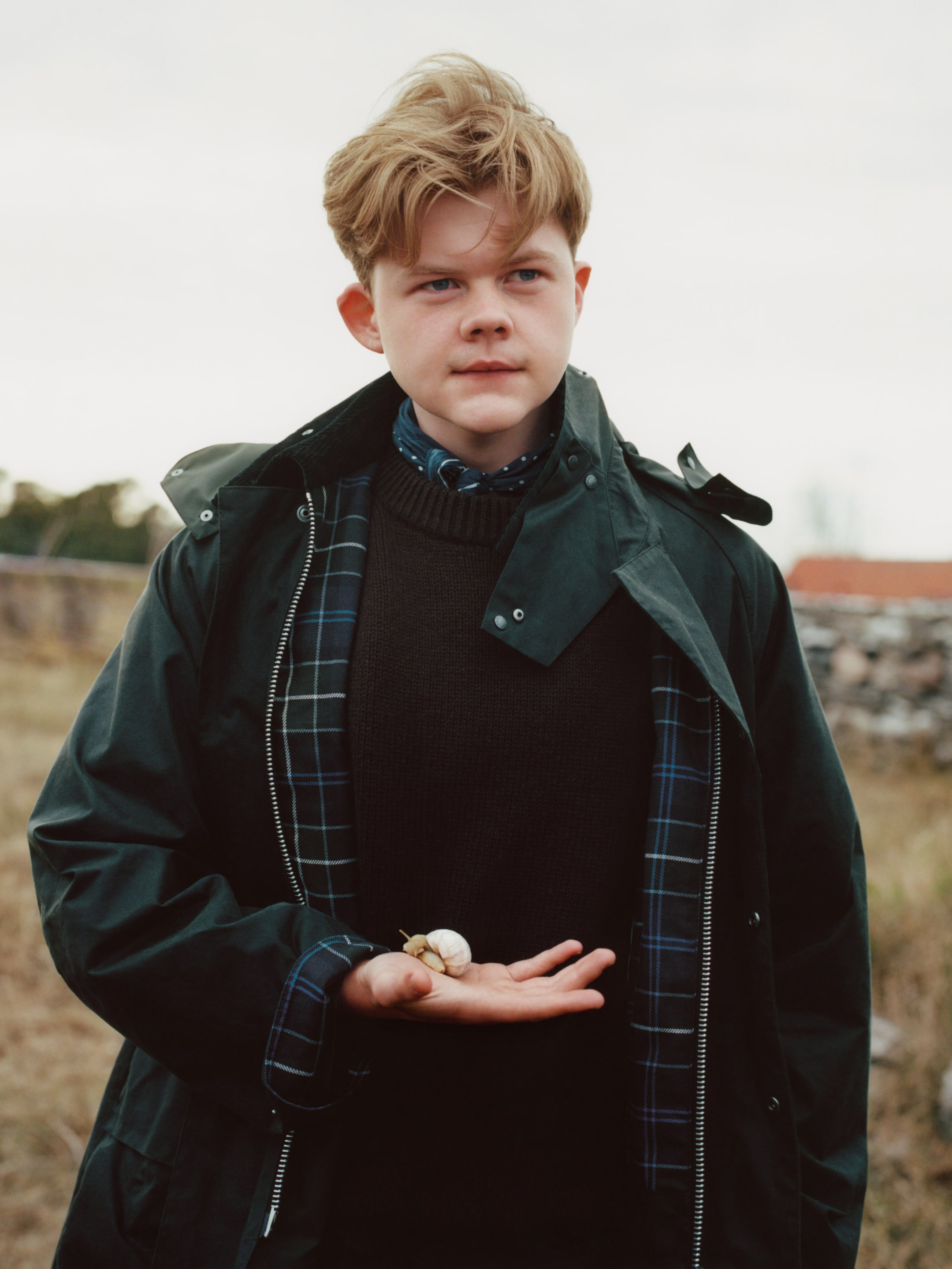 Young person with blond hair in a dark coat holds a small object in their palm, standing outdoors on a cloudy day.