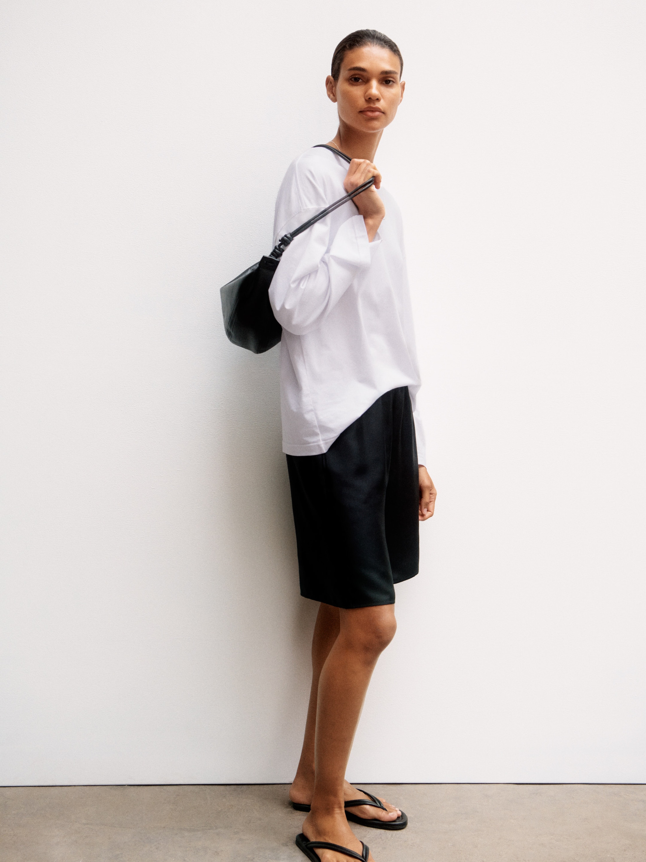 Person in a white long-sleeve shirt, black shorts, and flip-flops, holding a shoulder bag, standing against a white wall.