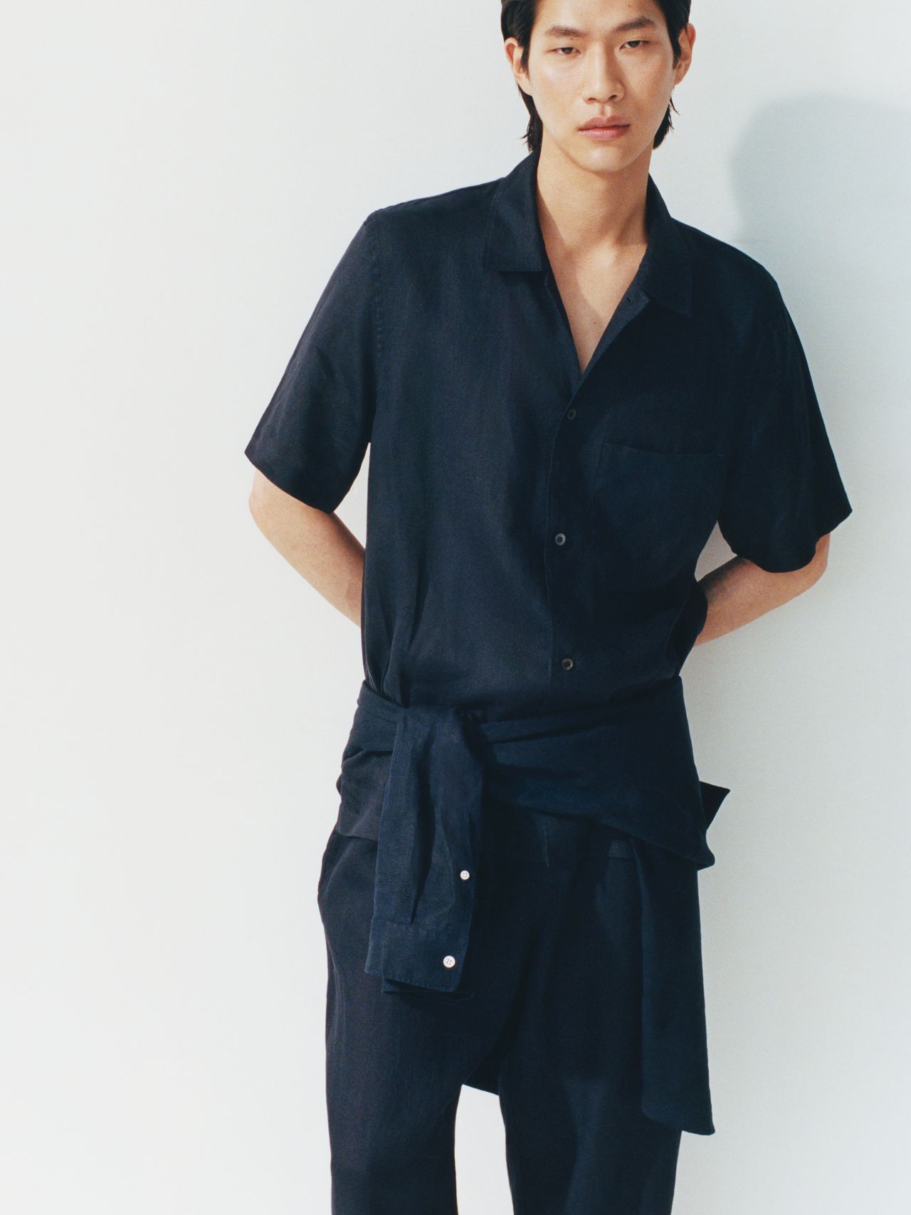Person wearing a dark short-sleeve shirt and pants, with a matching jacket tied at the waist, standing against a light background.