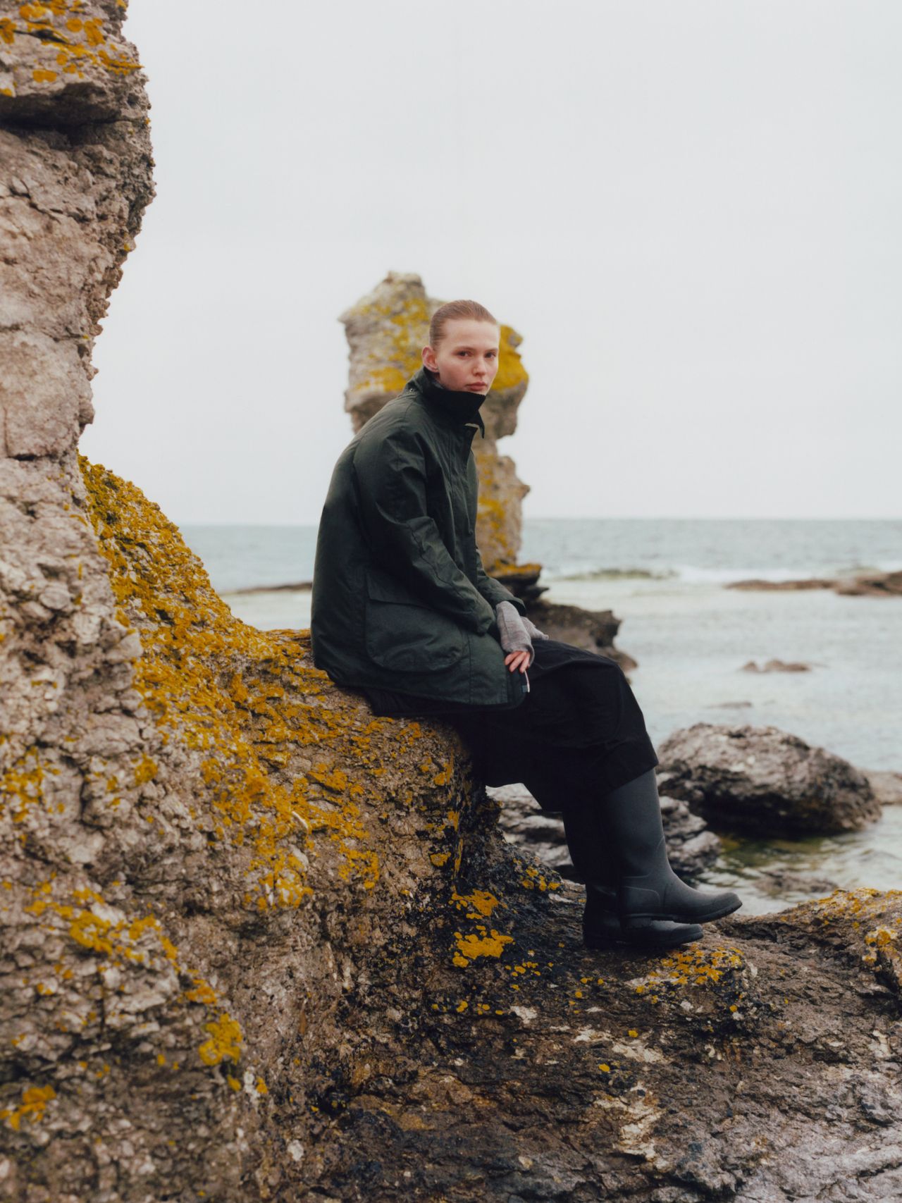 Person in dark clothing sits on a rocky shore with the ocean in the background, surrounded by lichen-covered rocks.