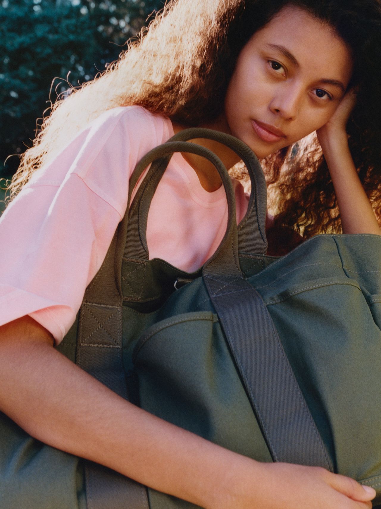 Person with long curly hair wearing a pink shirt, holding a large green bag, sitting outdoors with greenery in the background.