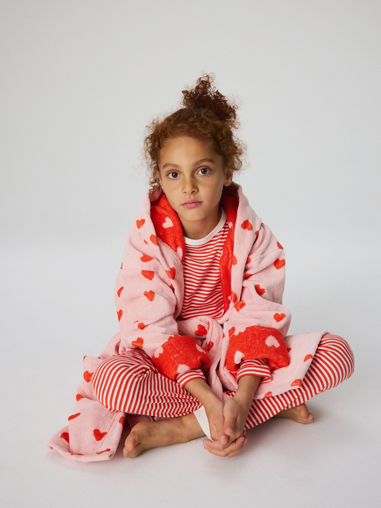 A child with curly hair sits cross-legged, wearing a pink robe with red hearts and red-and-white striped pajamas, against a plain background.