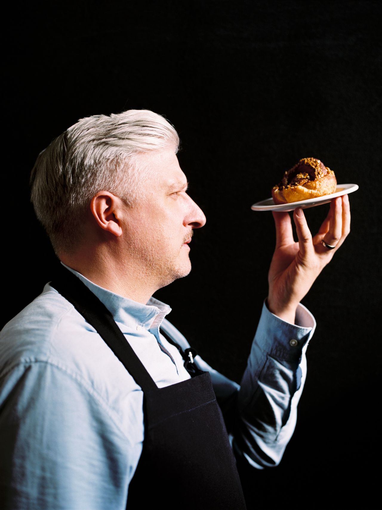 A man with gray hair and a blue shirt holds a pastry on a plate, gazing at it intently against a dark background.