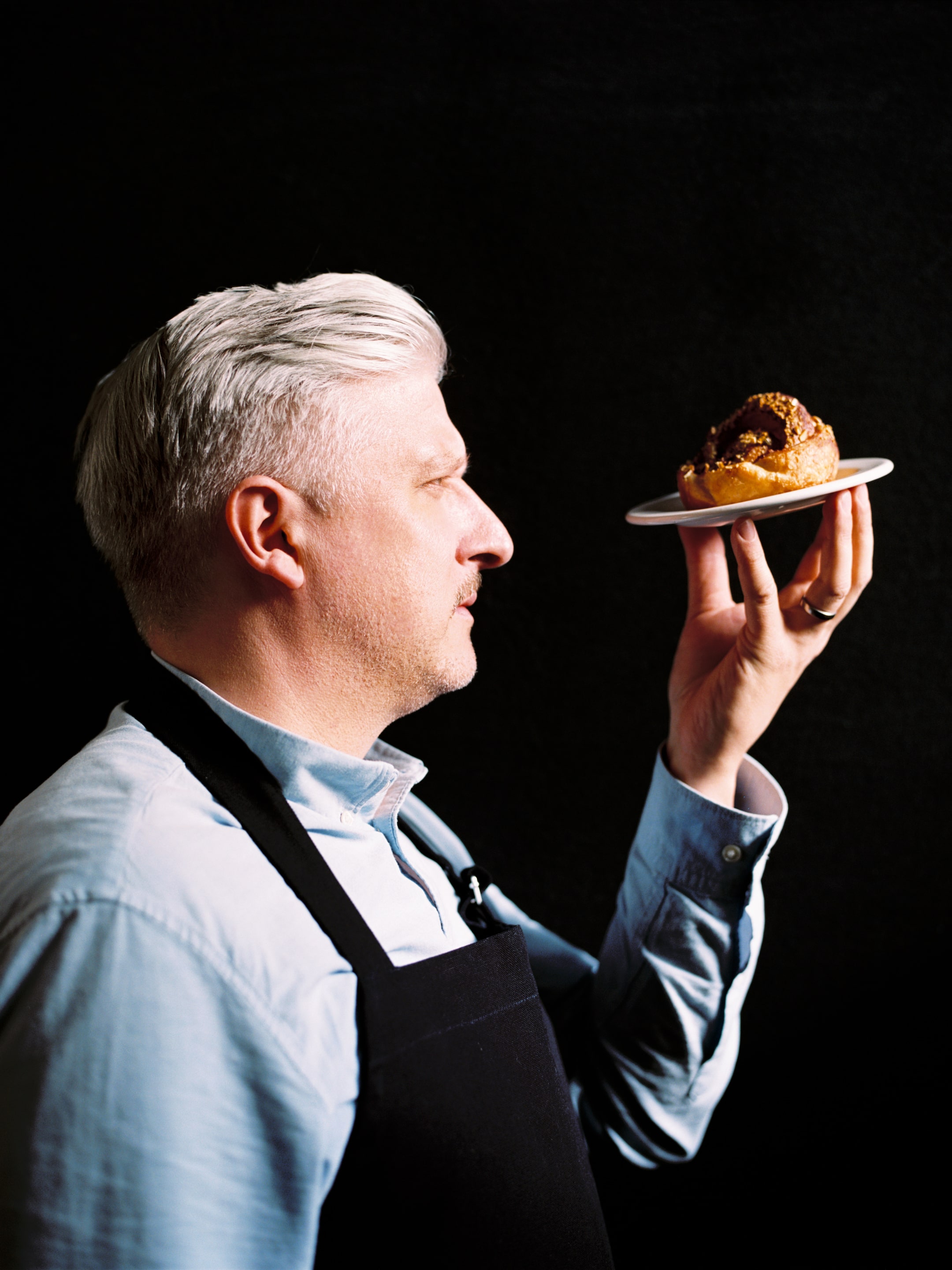 A man with gray hair and a blue shirt holds a pastry on a plate, gazing at it intently against a dark background.