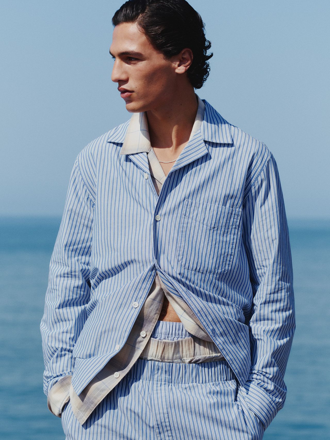 Man in a blue and white striped outfit stands by the sea, hands in pockets, gazing to the side against a clear blue sky.