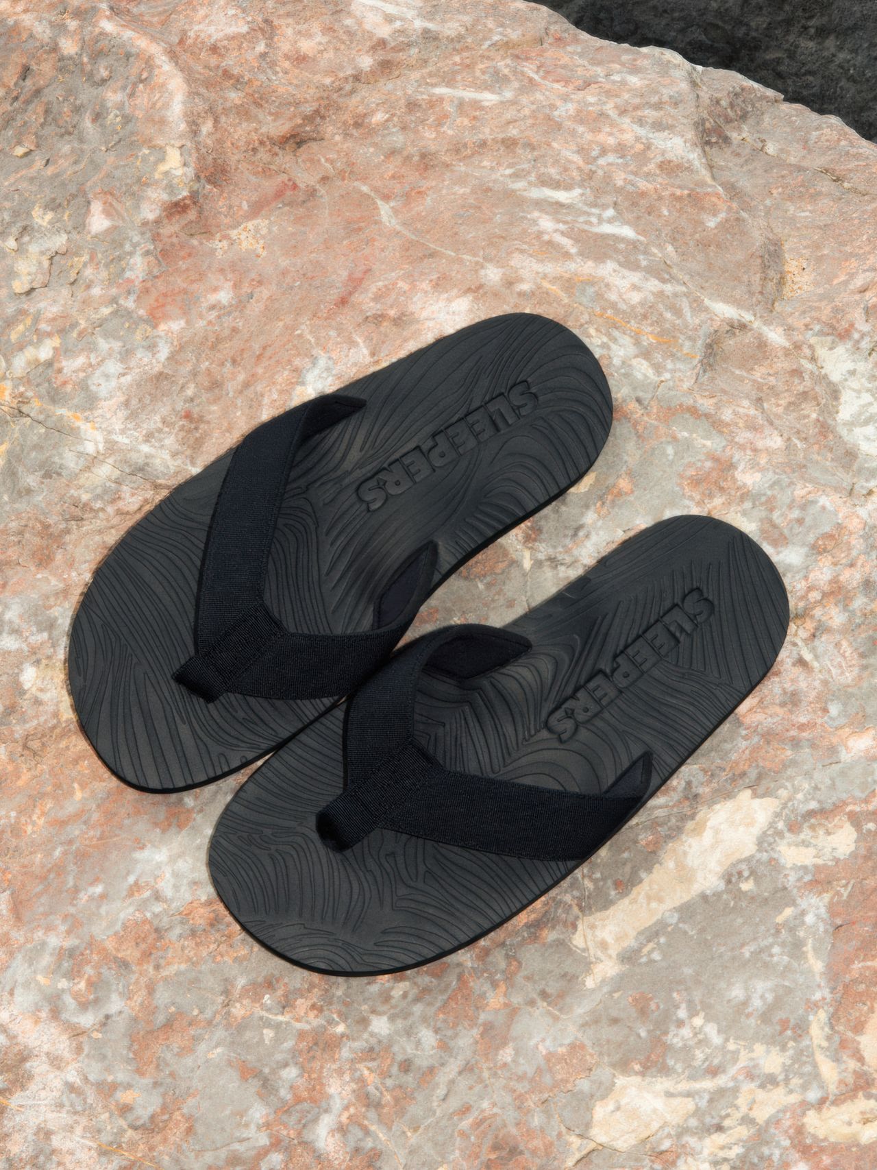 Black flip-flops with textured soles and "SLEEPERS" branding, placed on a rough, textured stone surface.