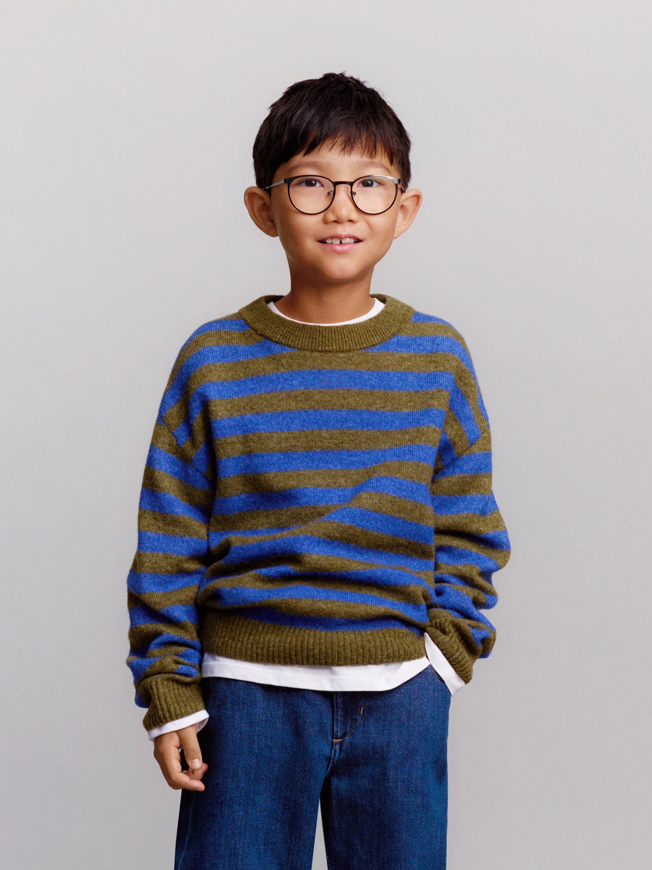 A young boy wearing glasses, a blue and olive striped sweater, and jeans stands smiling against a plain background.