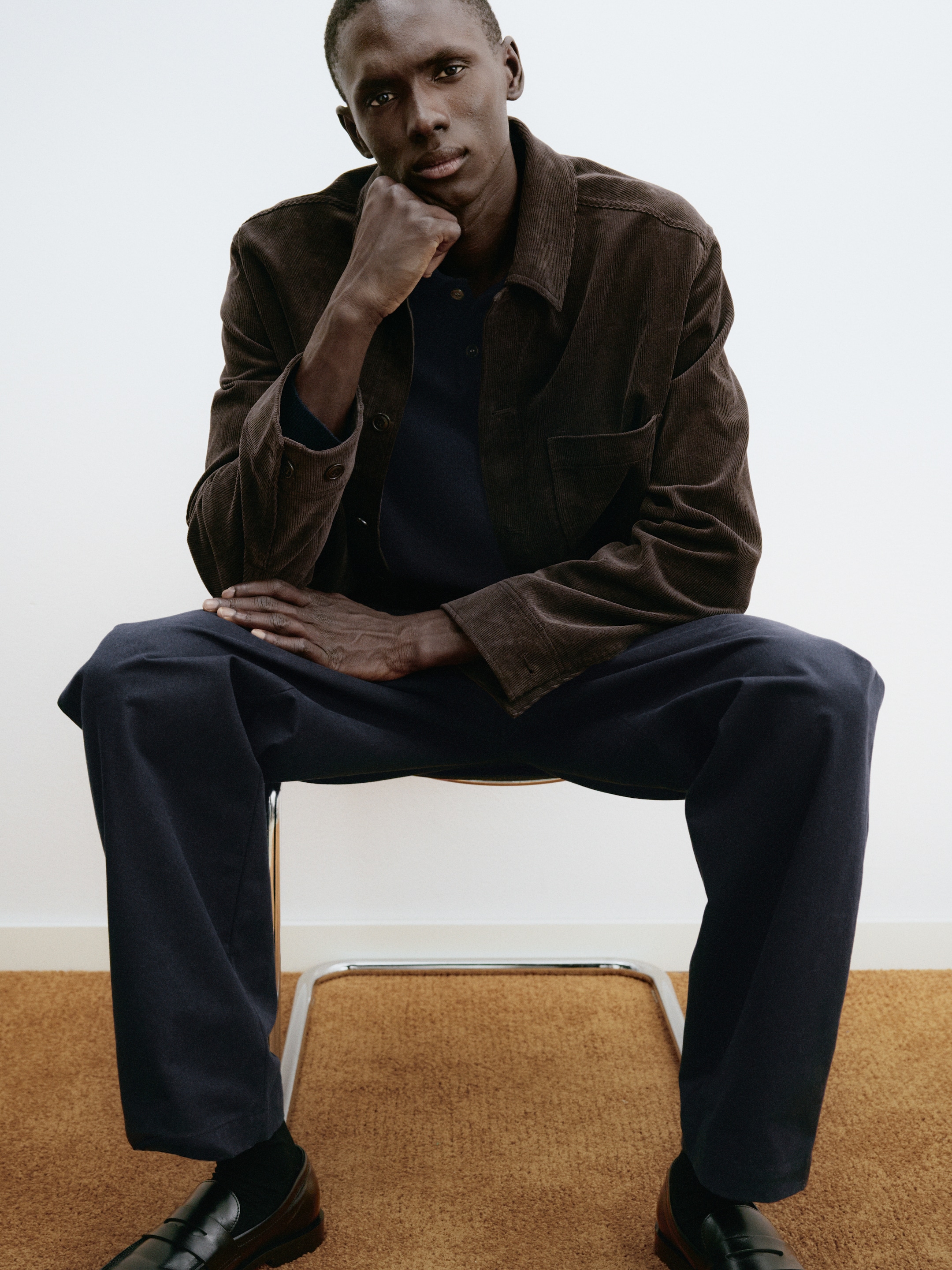 A person seated on a chair, wearing a brown jacket, navy pants, and black shoes, with one hand resting under their chin, looking thoughtful.