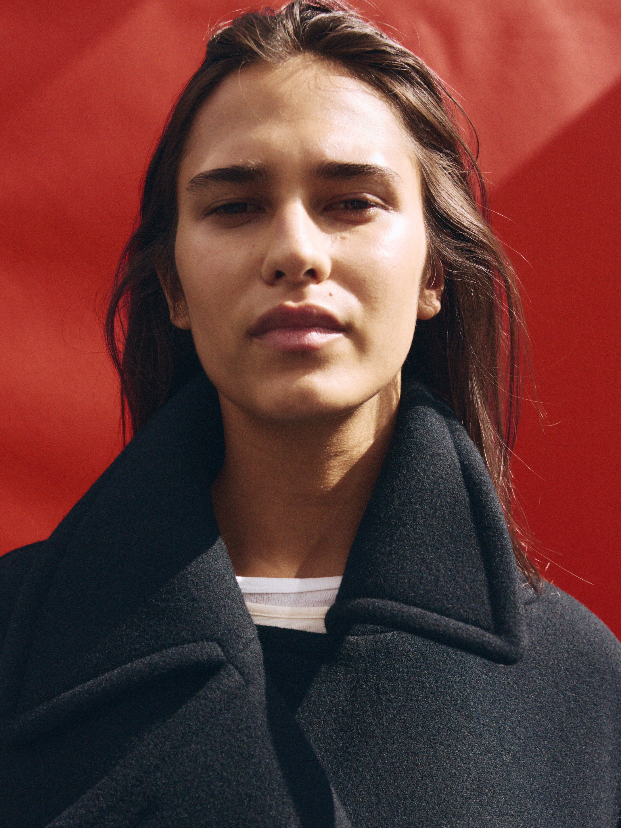 Person with long hair wearing a dark coat, standing in front of a vibrant red background, looking directly at the camera.