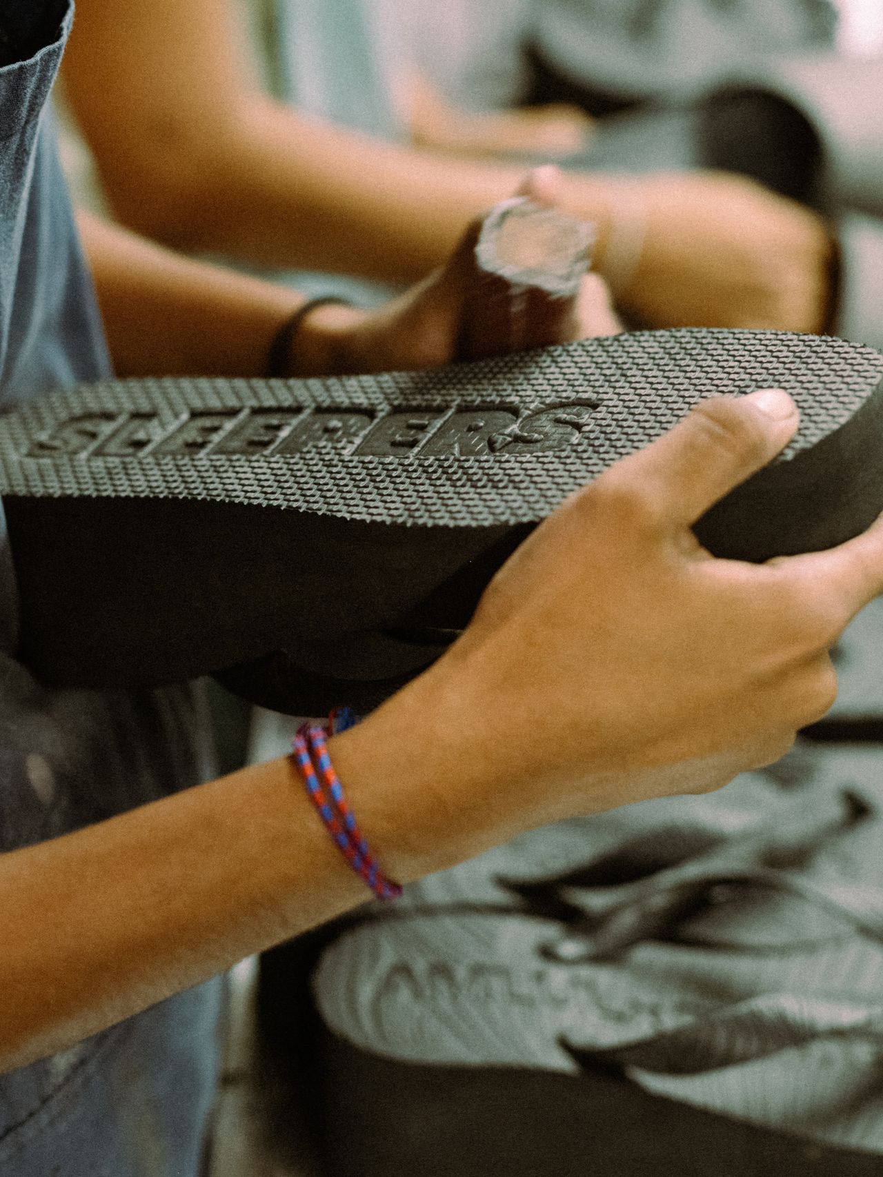Person holding a textured black sandal sole with the word "SLEEPERS" embossed, wearing a blue and red braided bracelet.