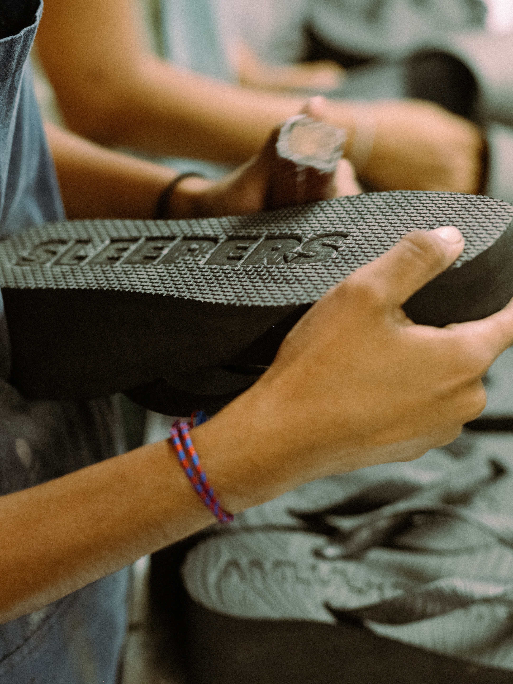 Person holding a textured black sandal sole with the word "SLEEPERS" embossed, wearing a blue and red braided bracelet.