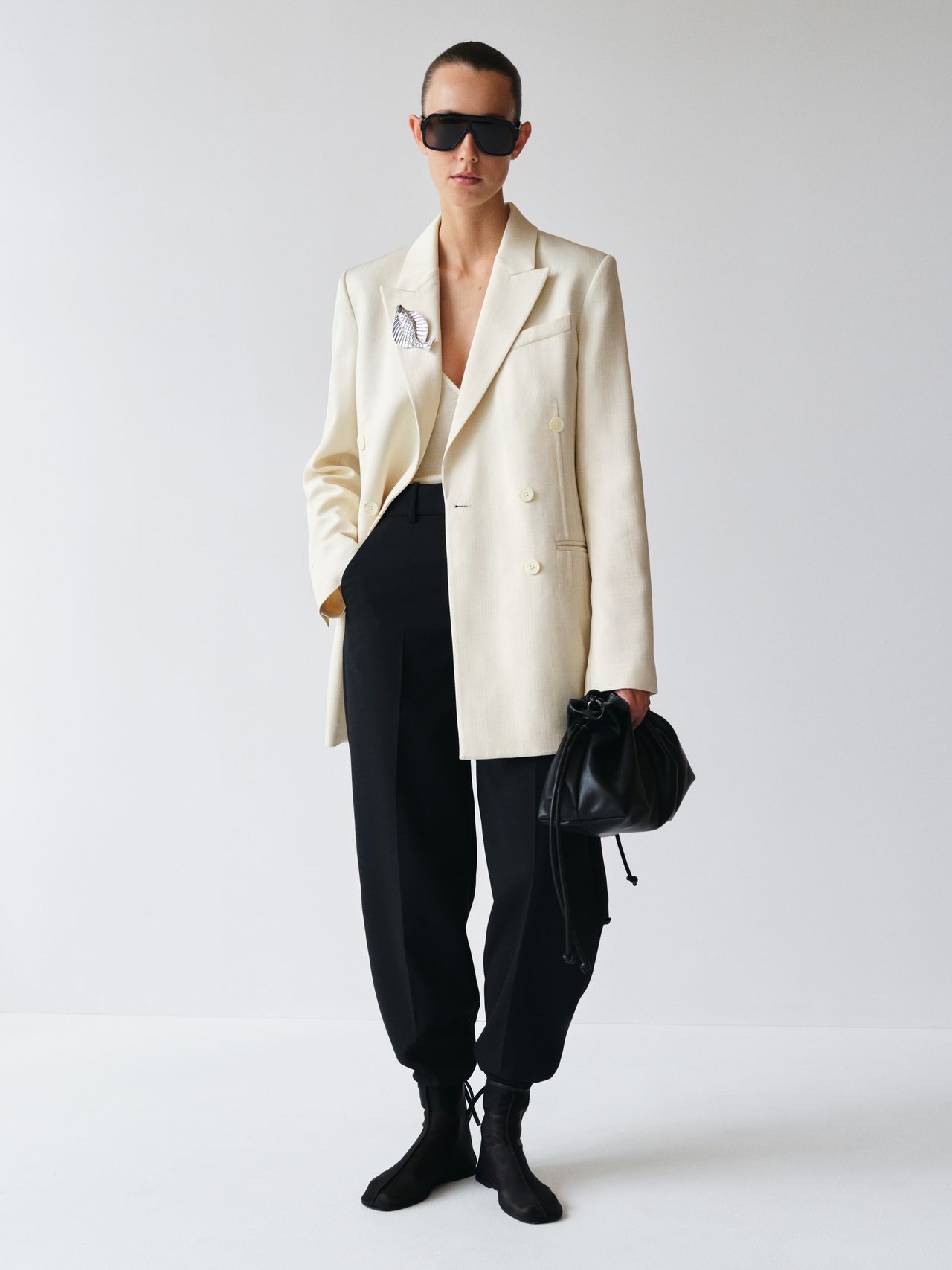 Person wearing a cream blazer, black pants, and sunglasses, holding a black bag, standing against a plain white background.