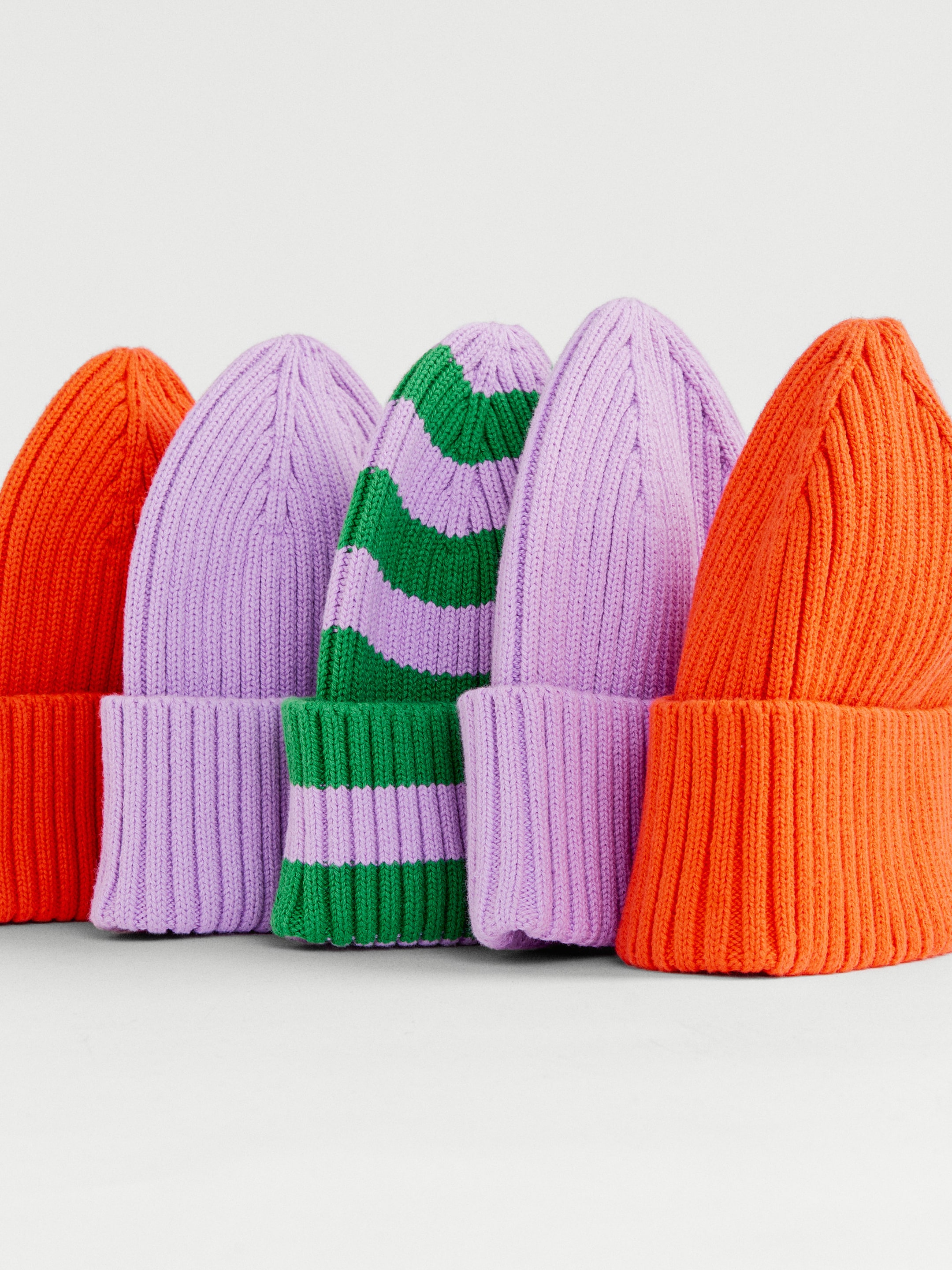 Five knit beanies arranged upright: two orange, two lavender, and one striped green and lavender.