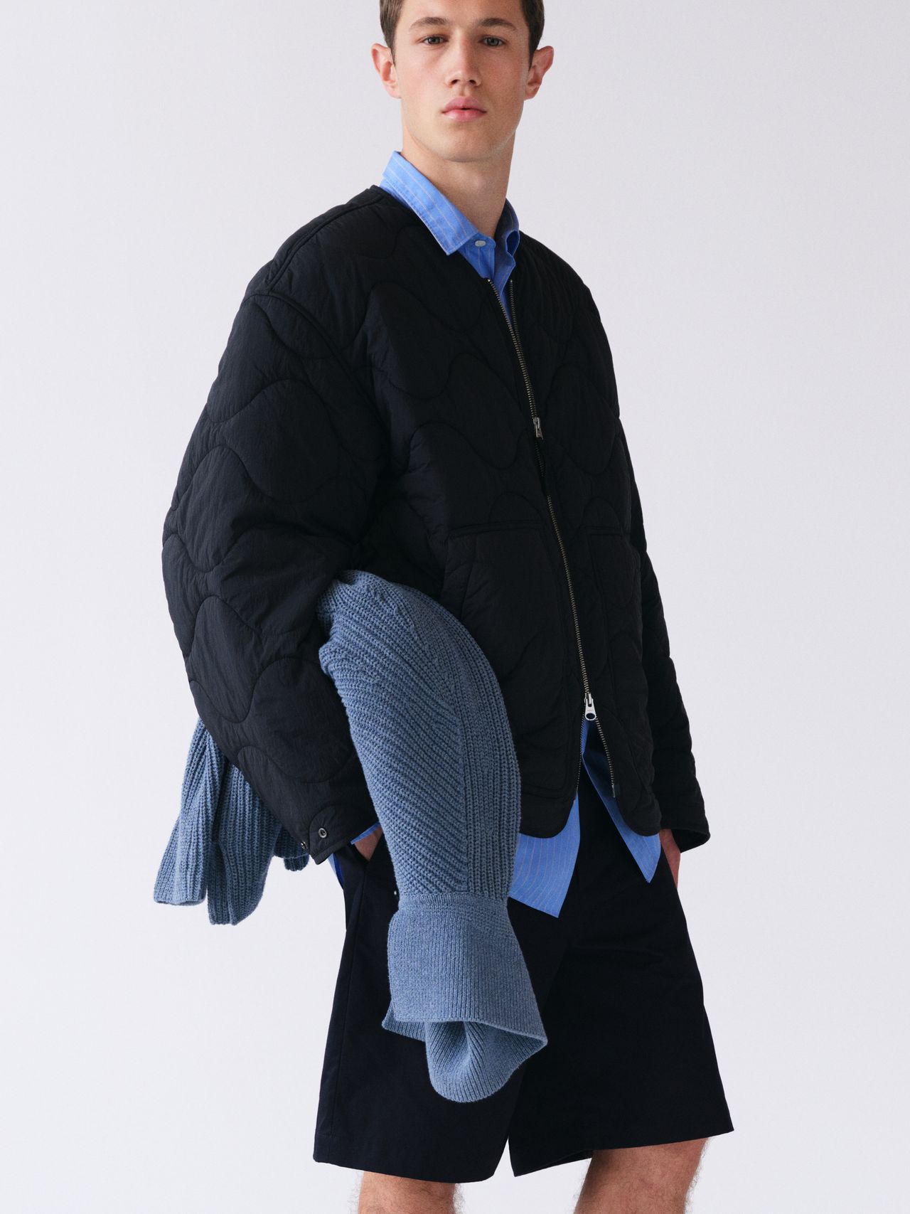 Young man in a black quilted jacket over a blue shirt, holding a blue knit sweater, wearing dark shorts, standing against a plain background.