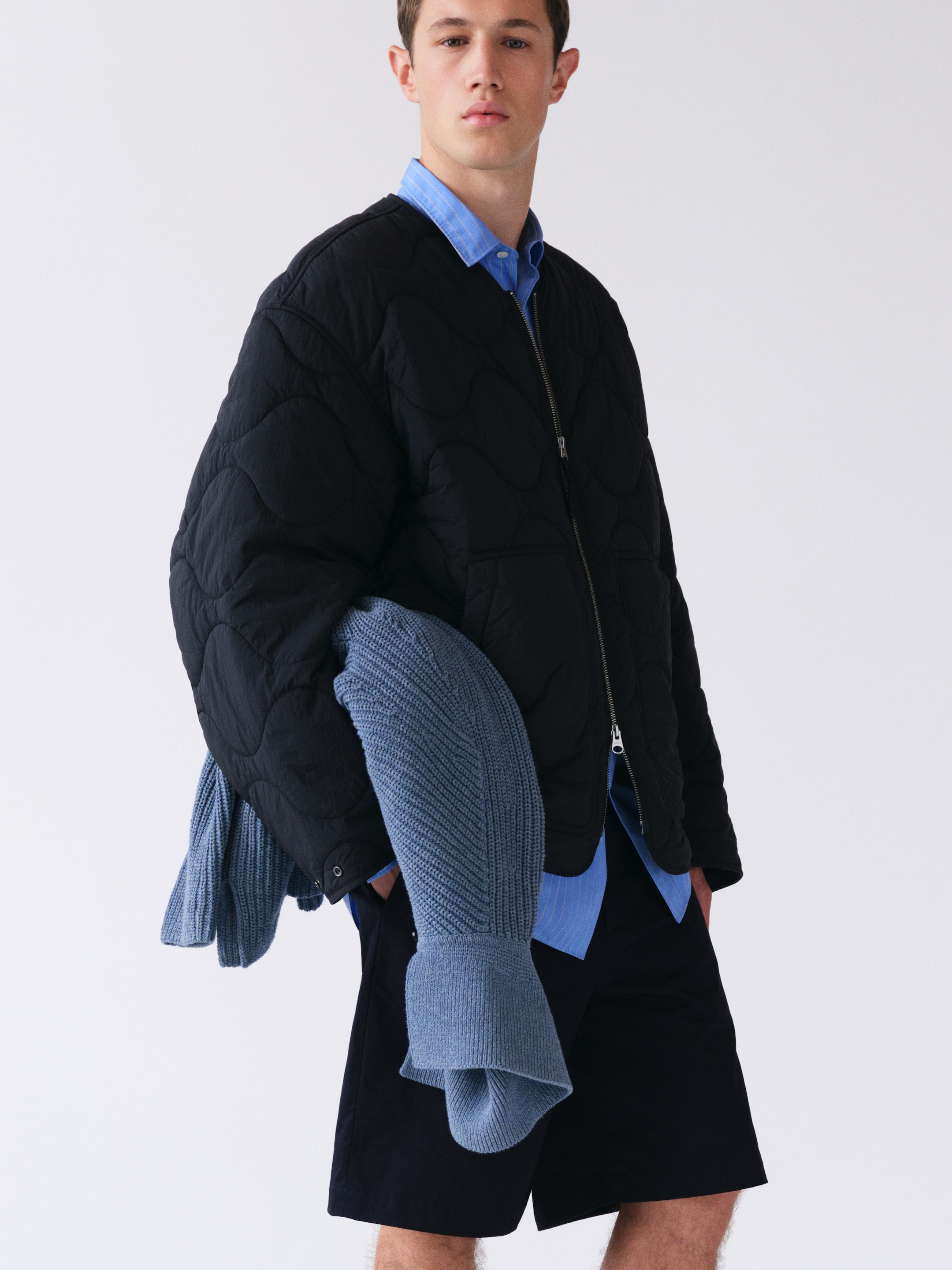 Young man in a black quilted jacket over a blue shirt, holding a blue knit sweater, wearing dark shorts, standing against a plain background.