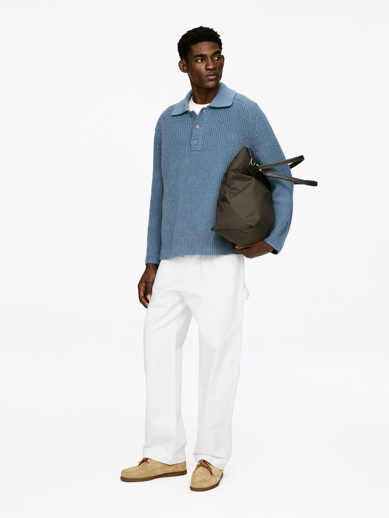 Man in a blue sweater and white pants holding a gray bag, standing against a plain white background.