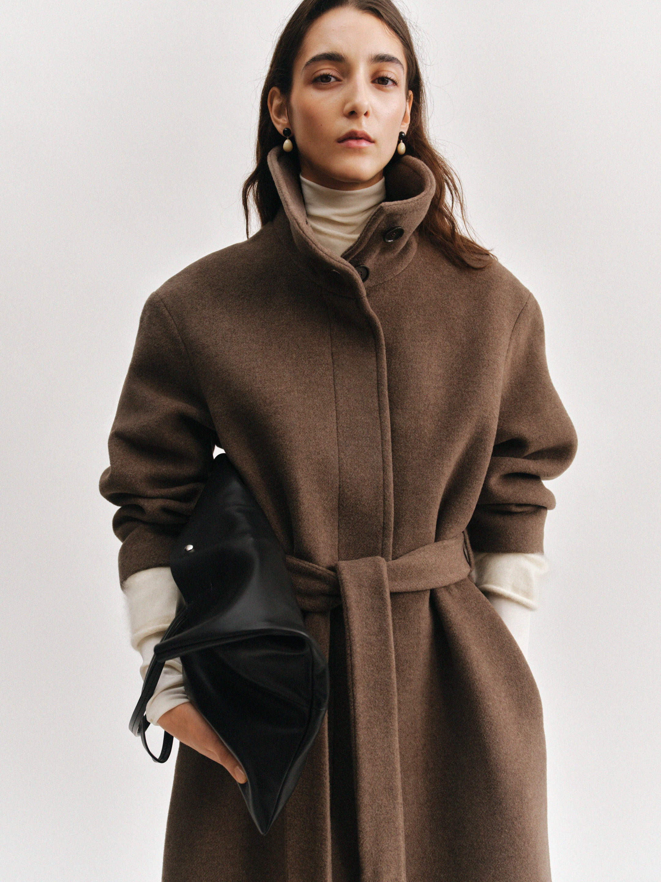 Woman in a tan coat with a belt and turtleneck underneath, holding a black clutch, wearing pearl drop earrings against a neutral background.