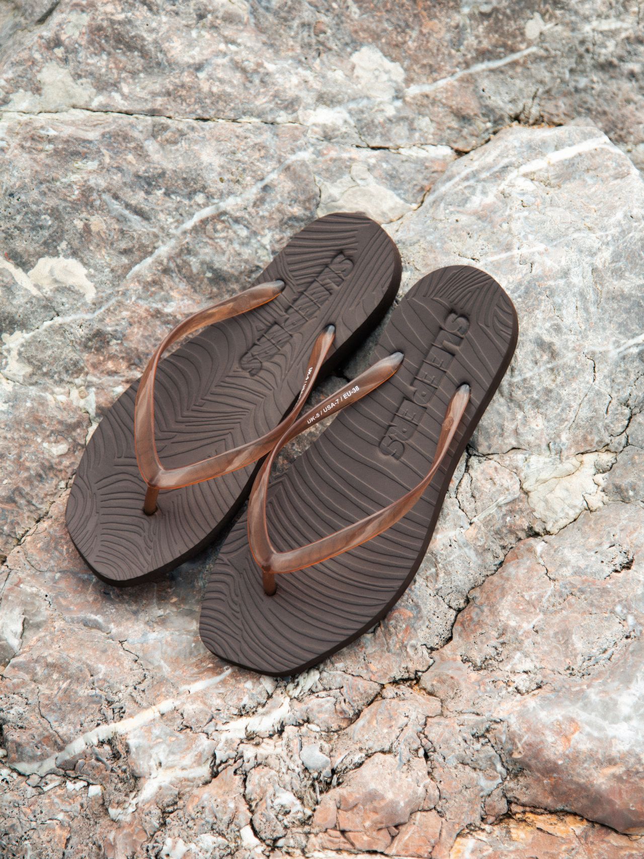 Brown flip-flops with wavy texture placed on a rough, rocky surface, showing the brand logo imprinted on the soles.