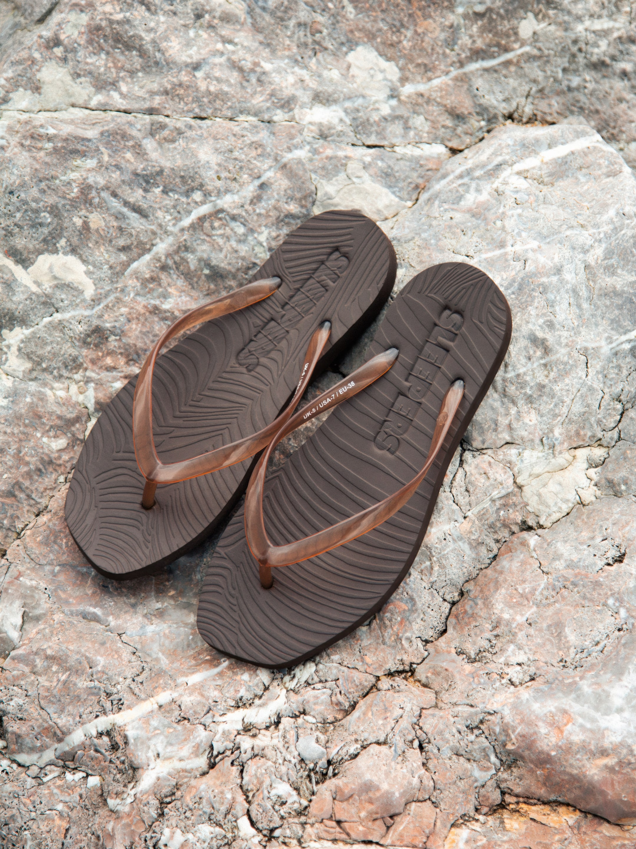 Brown flip-flops with wavy texture placed on a rough, rocky surface, showing the brand logo imprinted on the soles.