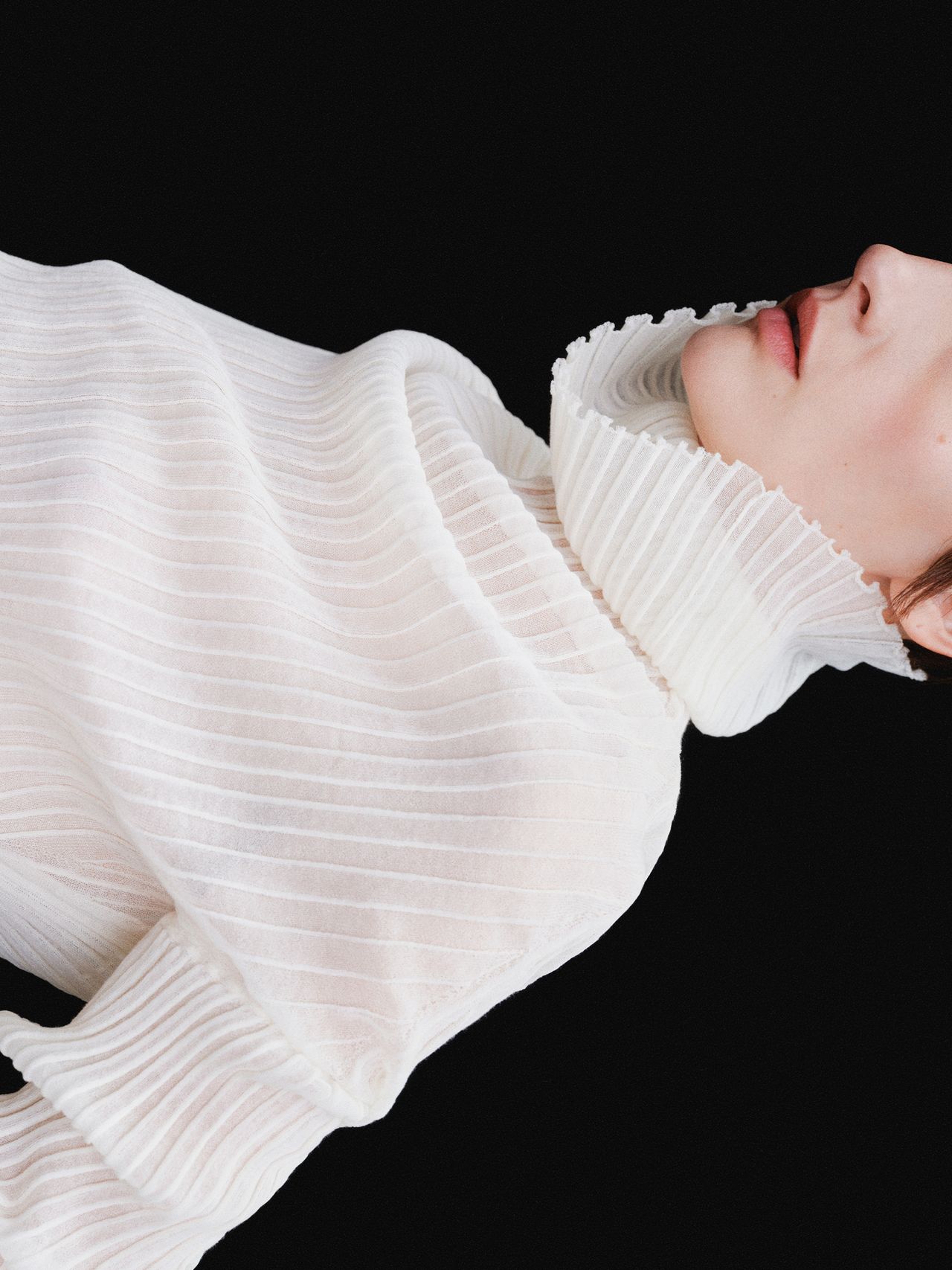 Person wearing a textured, white, high-collared garment against a black background, partially visible face.