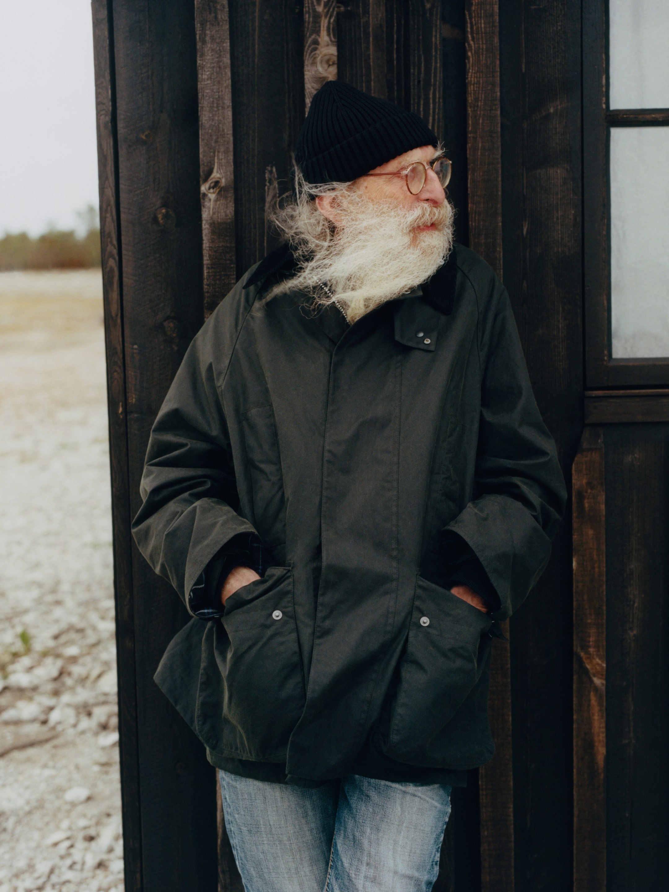 Elderly man with a long white beard, wearing a black beanie and coat, leans against a dark wooden wall, looking to the side.
