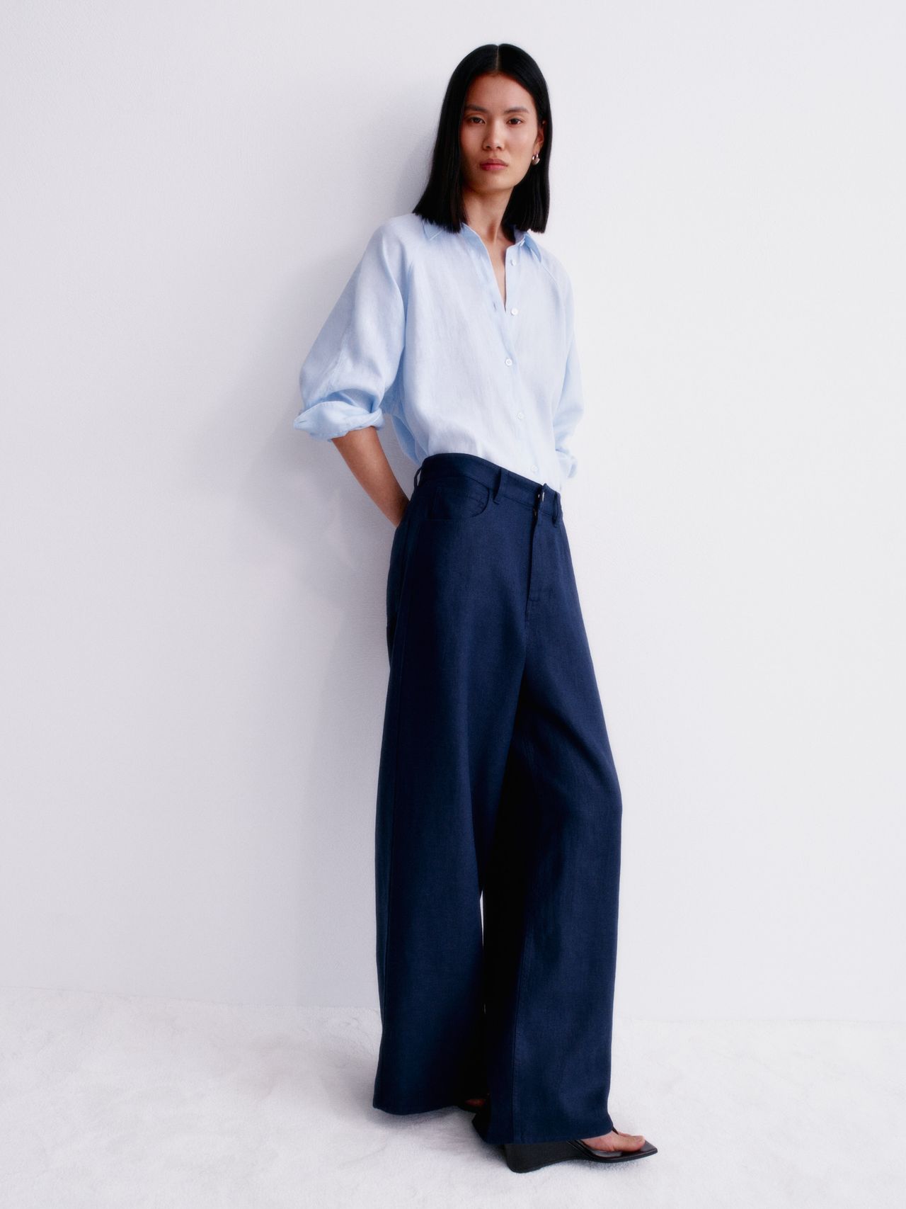 Person wearing a light blue shirt and wide-leg navy pants stands against a white wall, with arms relaxed by their sides.