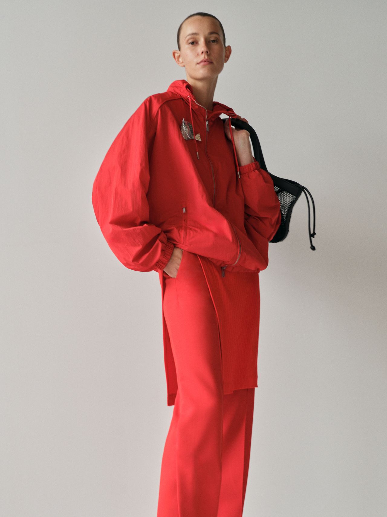 Person in a red outfit with a hooded jacket and pants, holding a black bag, standing against a plain background.