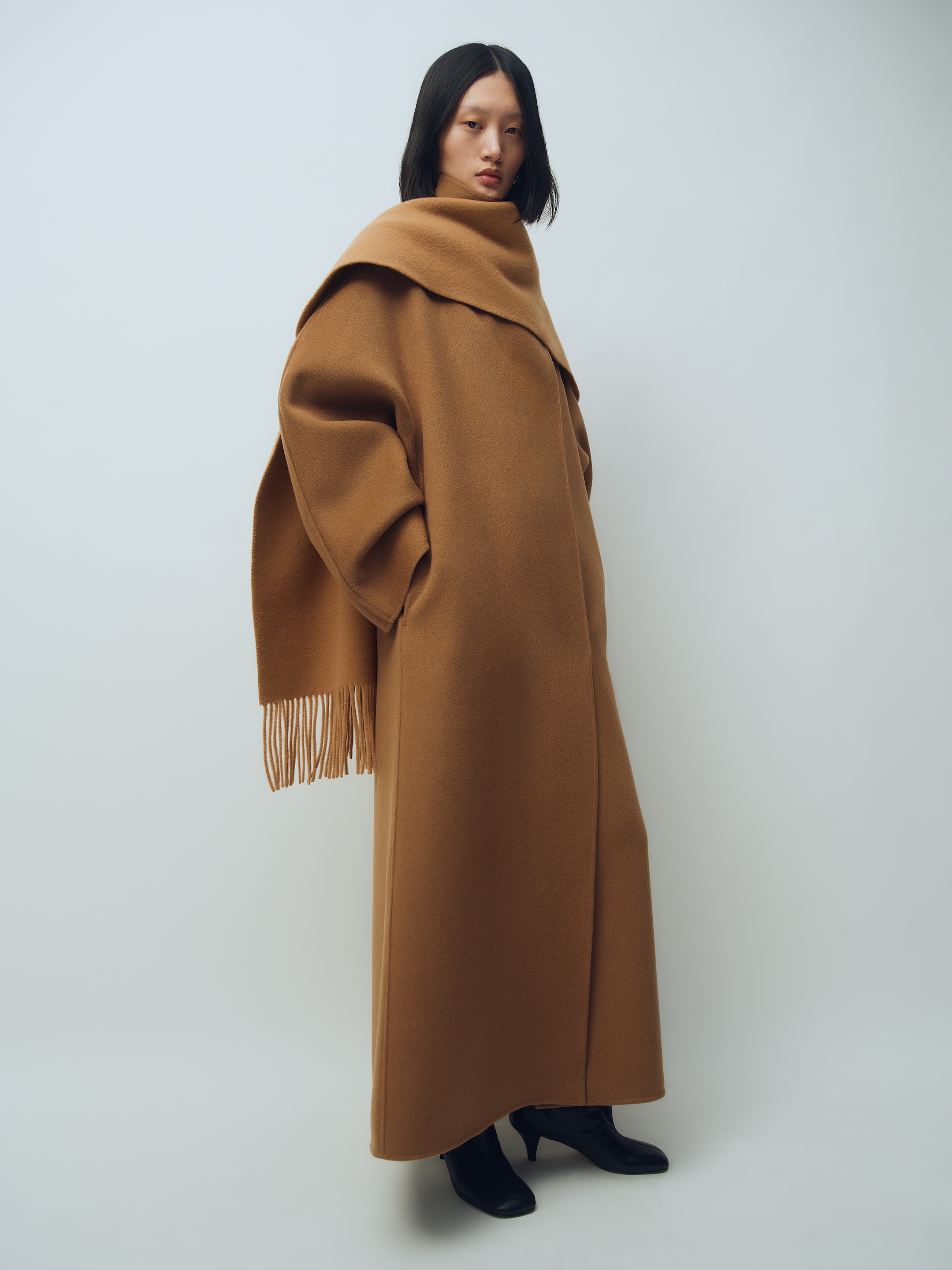 Person wearing a long brown coat with a fringed scarf, standing against a plain background in black heeled boots.