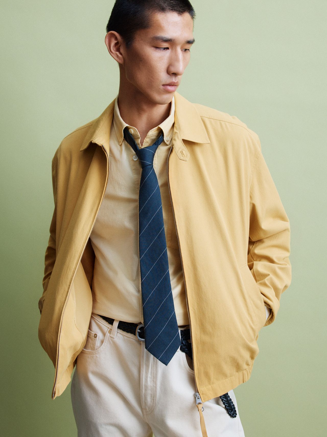 Person in a yellow jacket and shirt with a blue tie, standing against a green background, hands in pockets, looking sideways.