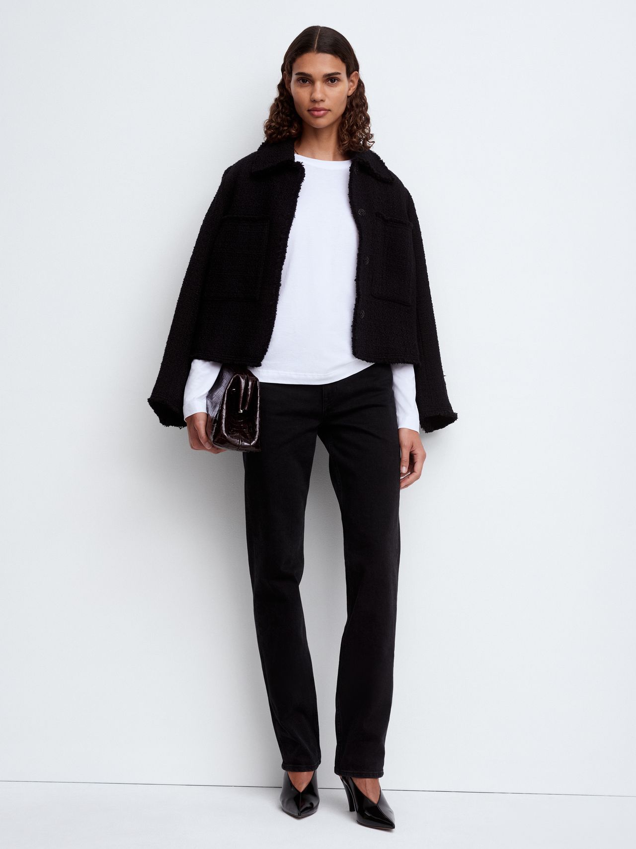 Woman in a black jacket, white shirt, and black pants, holding a clutch. She stands against a plain white background.