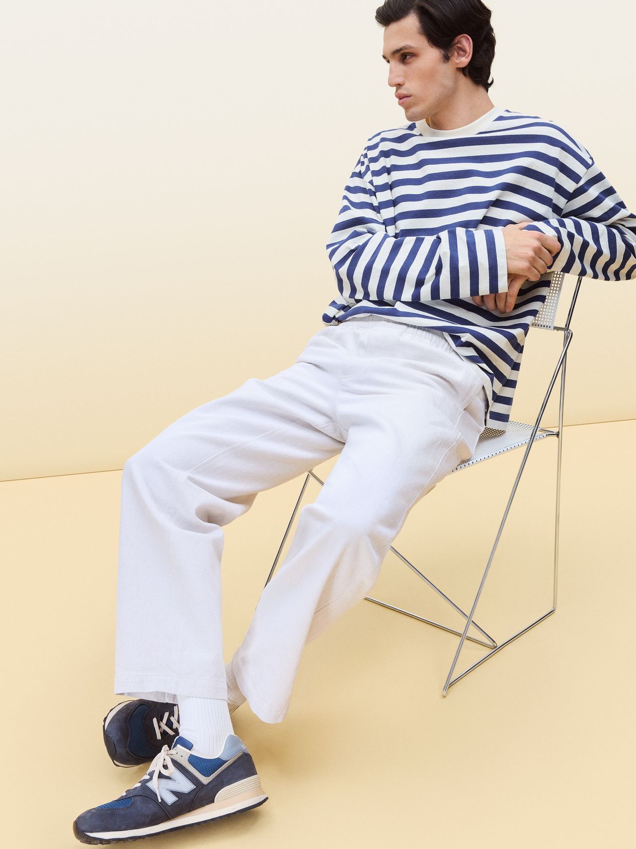 Man in a striped long-sleeve shirt and white pants sits on a wire chair, wearing blue sneakers against a beige background.