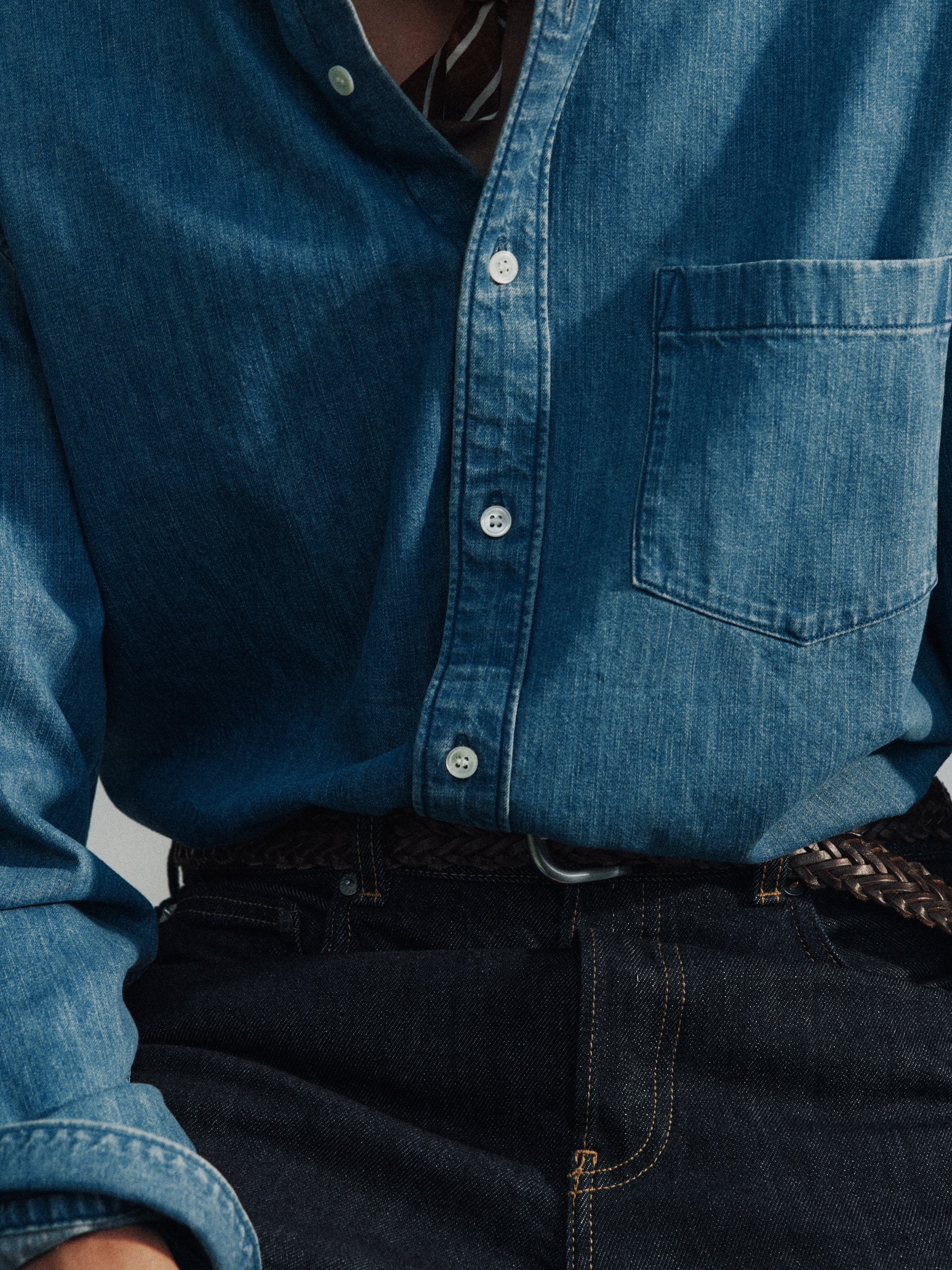 Close-up of a person wearing a blue denim shirt and dark jeans, featuring a braided leather belt.