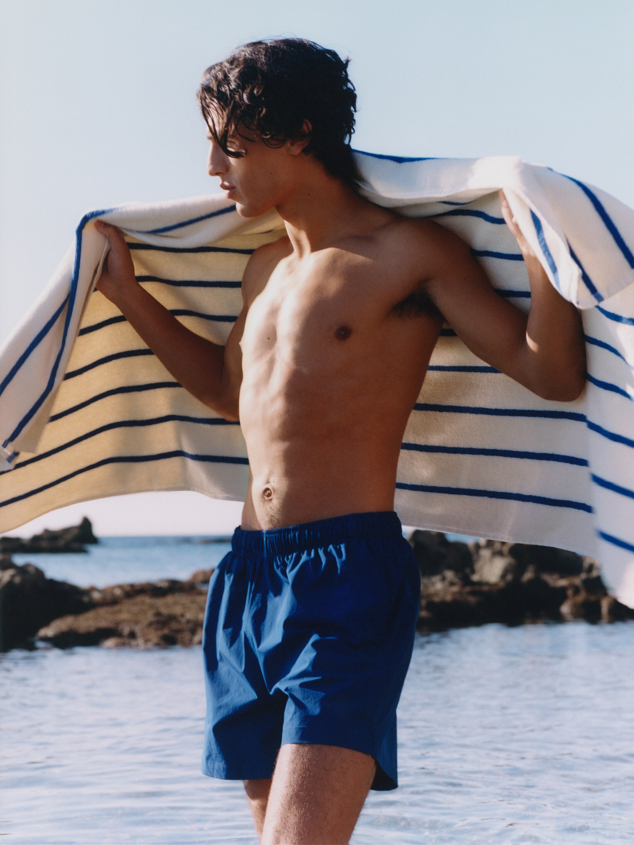 A person stands on a beach with a striped towel draped over their shoulders, wearing blue shorts. The ocean and rocks are visible in the background.