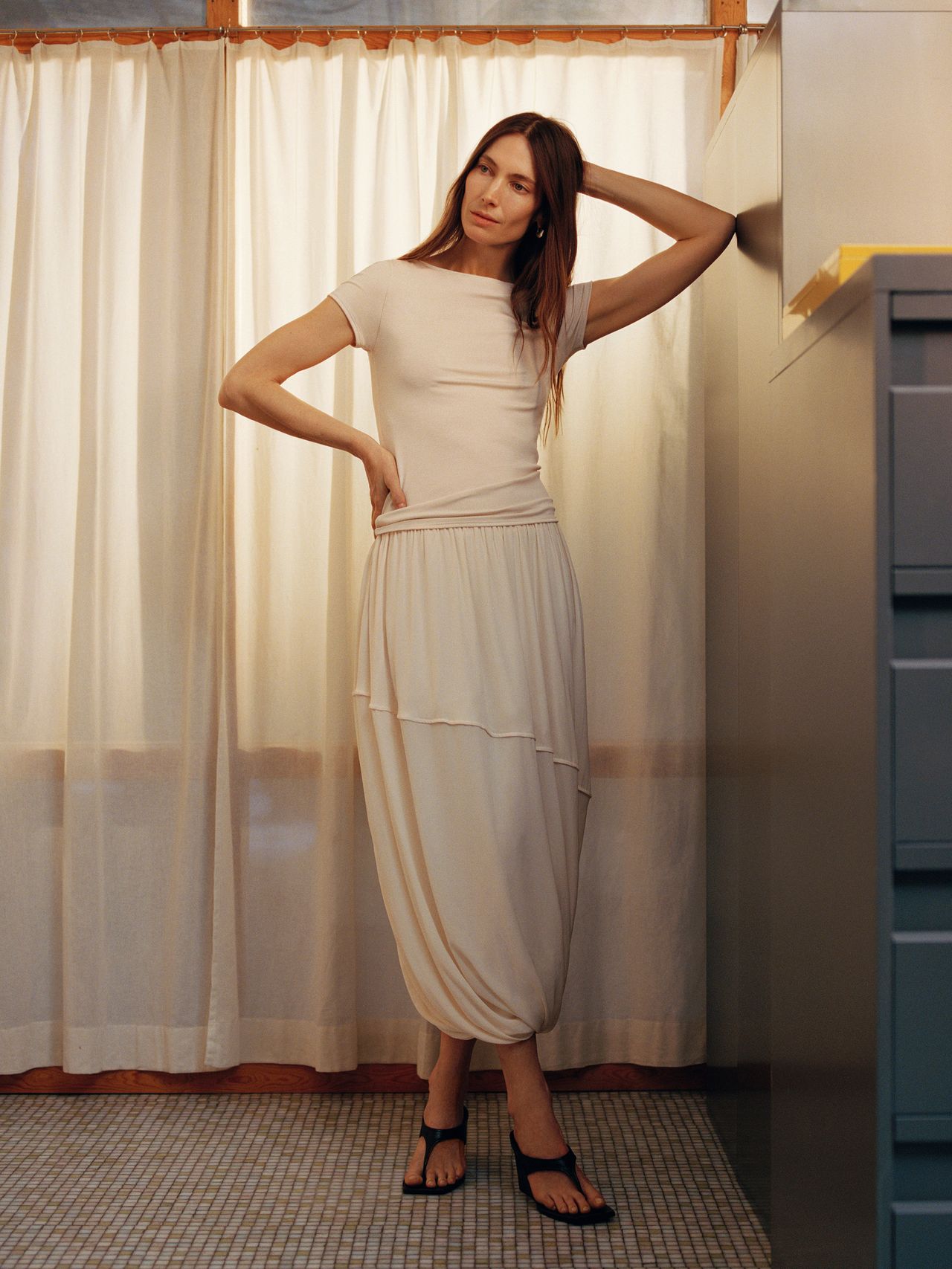 A woman in a white dress and sandals stands in a room with beige curtains, posing with one hand on her head and the other on her hip.